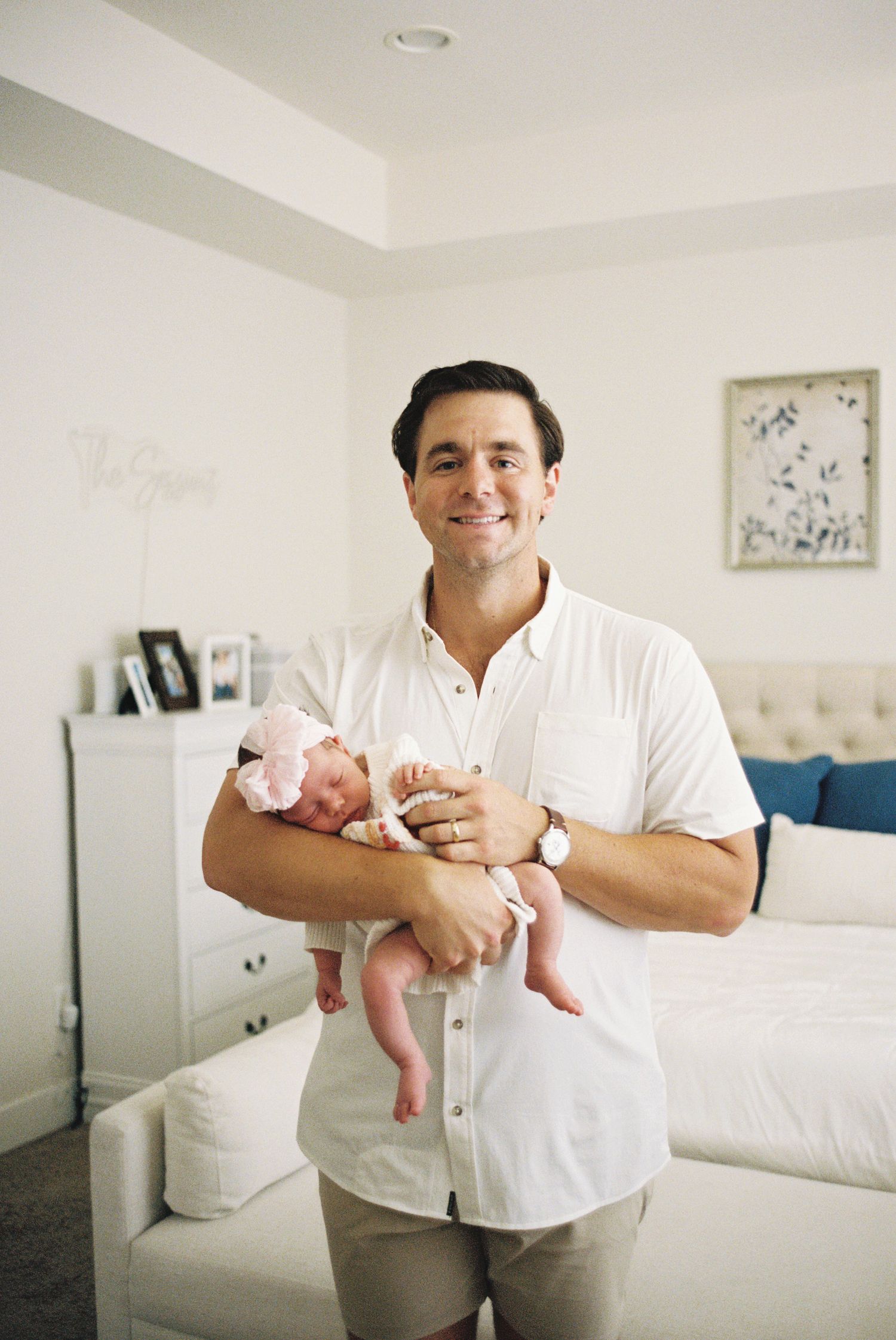 Smiling man in white shirt holding a sleeping newborn baby in pink outfit inside a bright modern bedroom.
