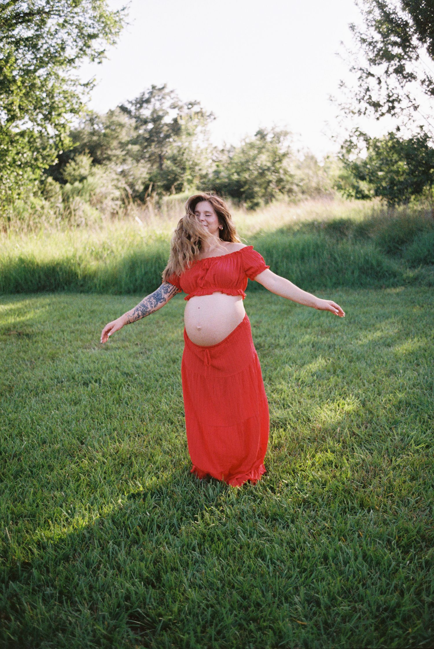 Pregnant woman in red off-shoulder dress standing in green field with arms outstretched, showing baby bump.