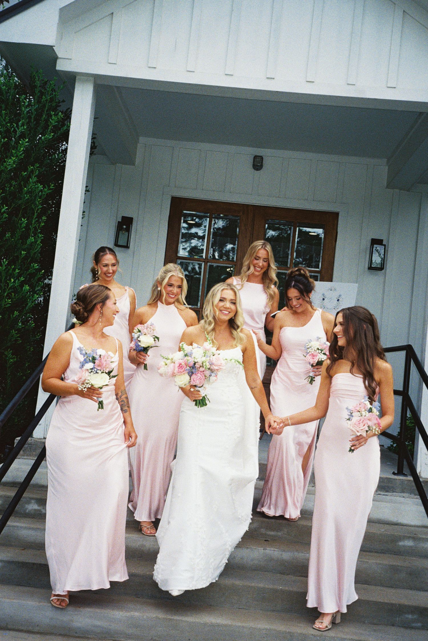 Bride in white gown walks with six bridesmaids in blush pink dresses, holding bouquets on venue steps.