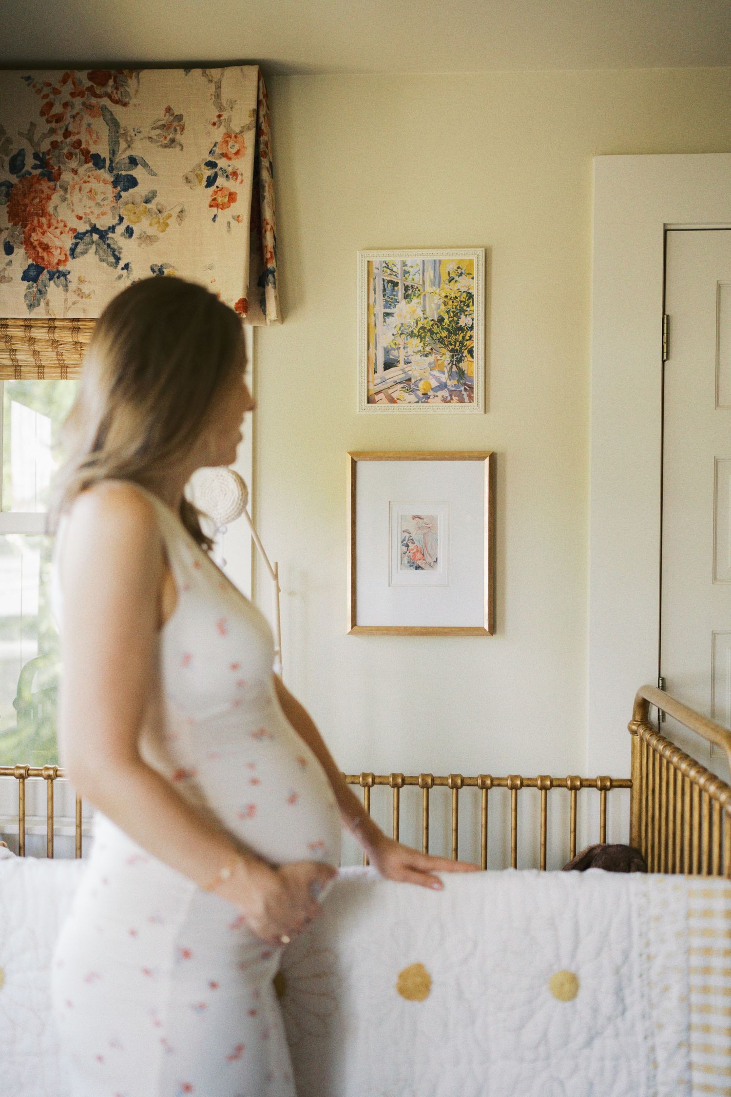 Pregnant woman in floral dress stands by gold crib in soft nursery with framed art and floral roman shade.