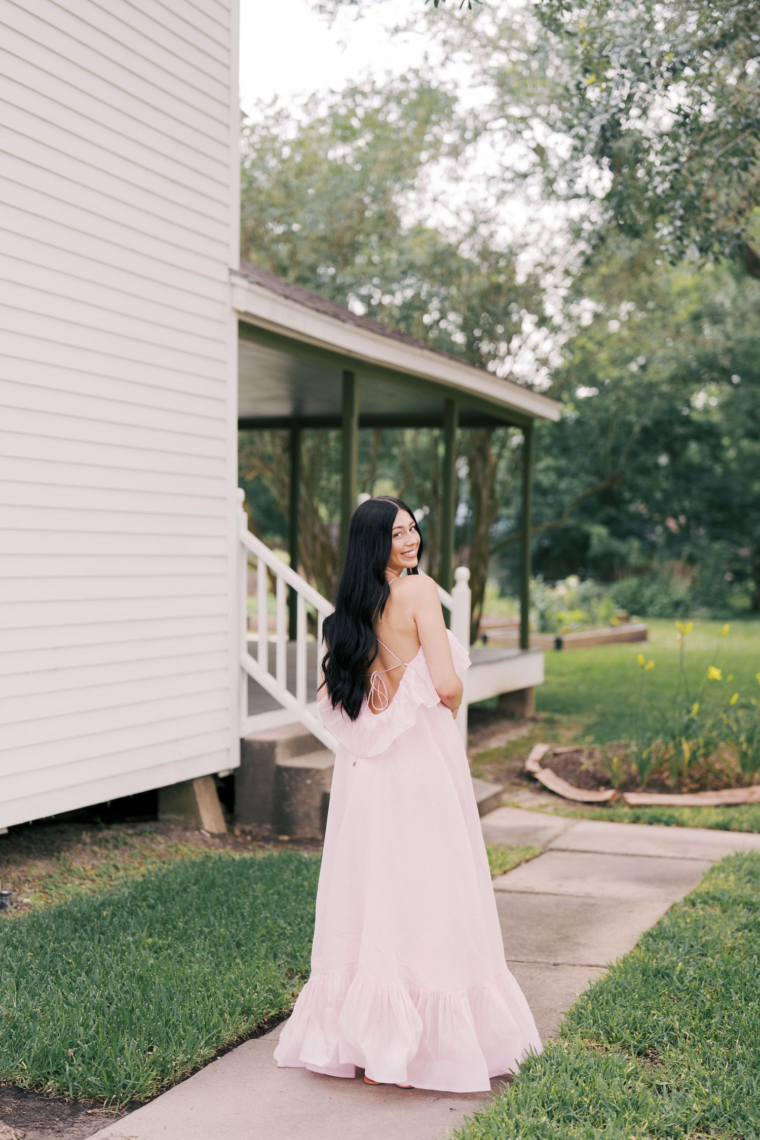 A woman in a pink maxi dress standing near a white porch with trees in the background.