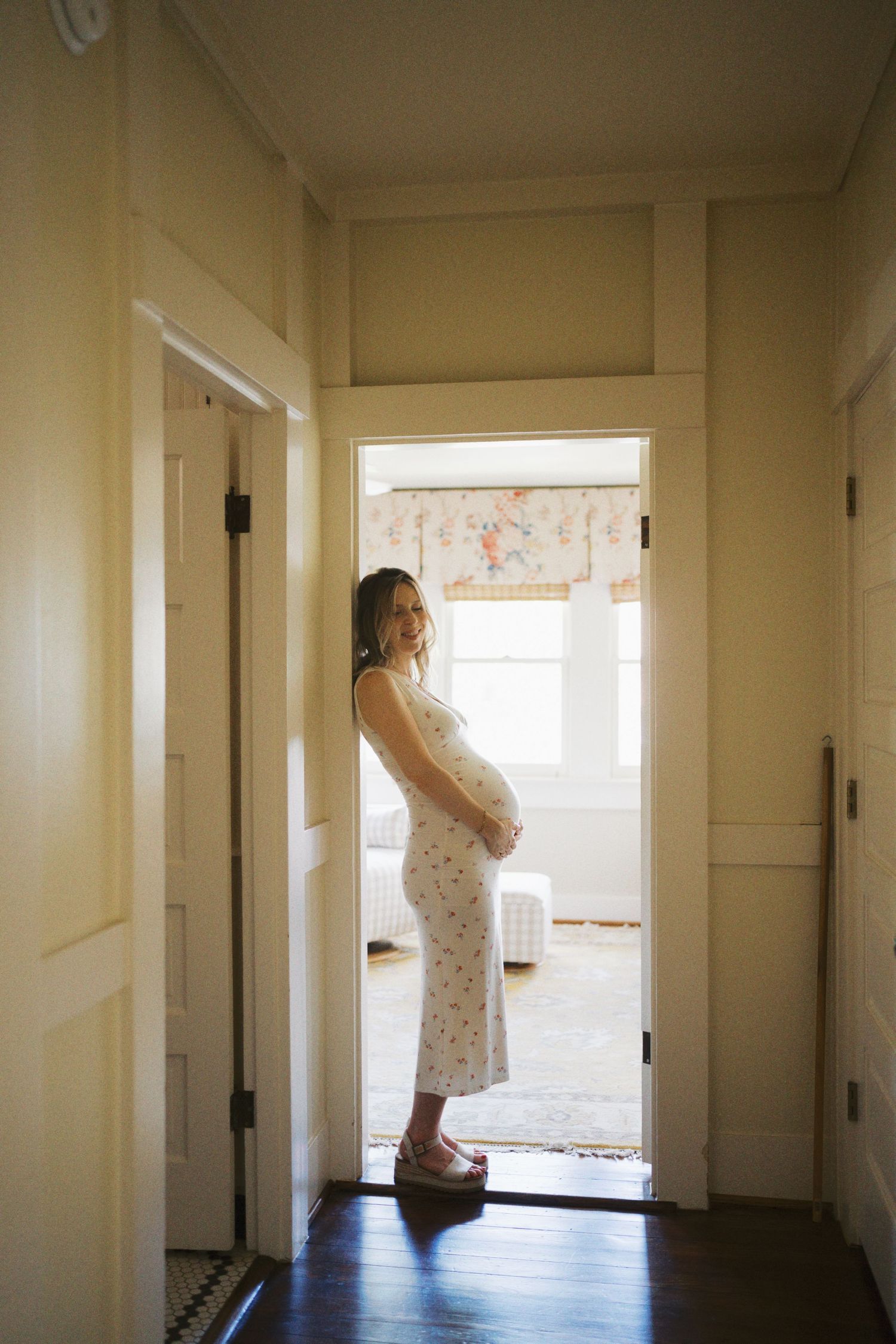 Pregnant woman in floral dress cradling baby bump in sunlit hallway doorway.