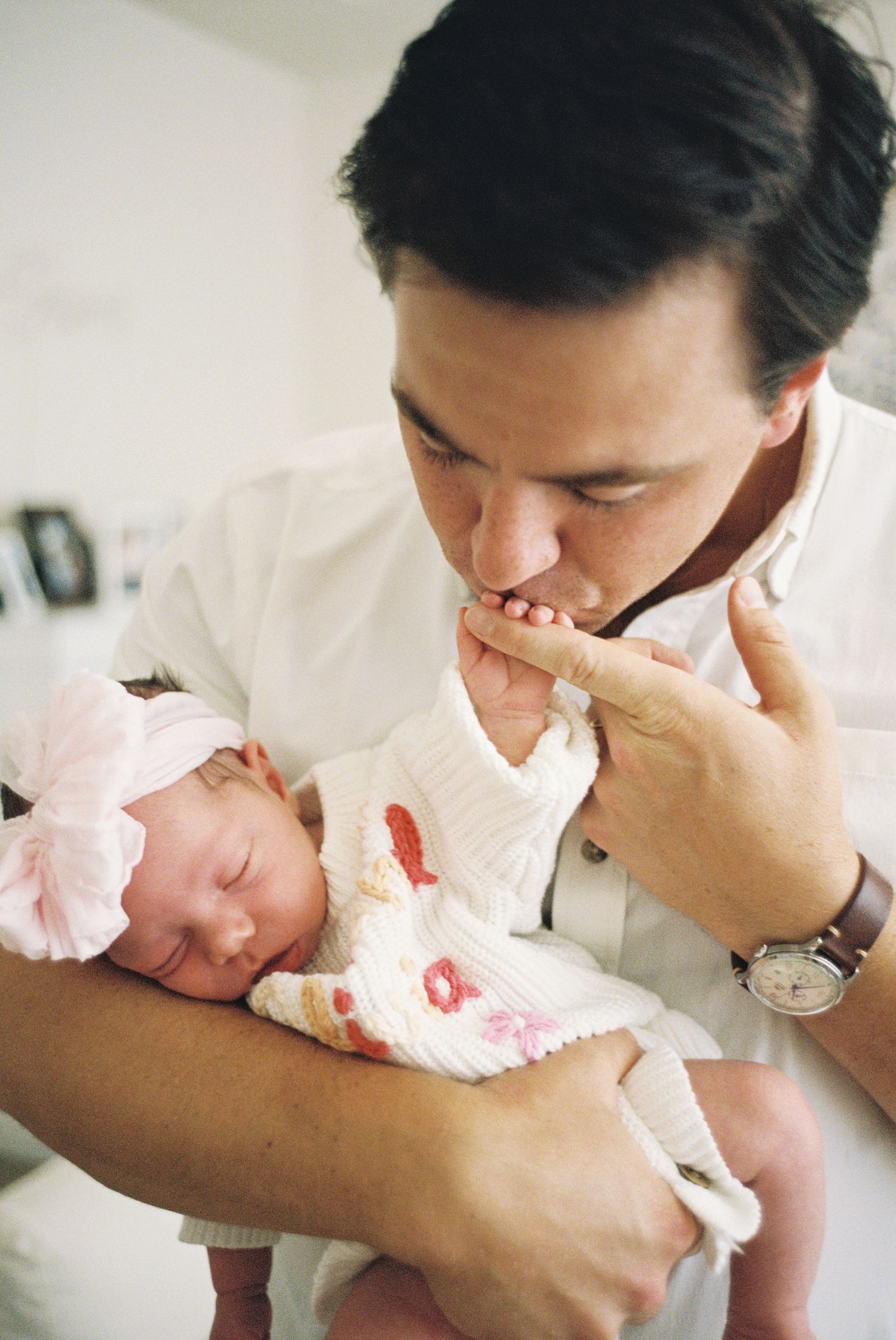 A father tenderly kisses his newborn baby's tiny hand while cradling her in a hospital room.