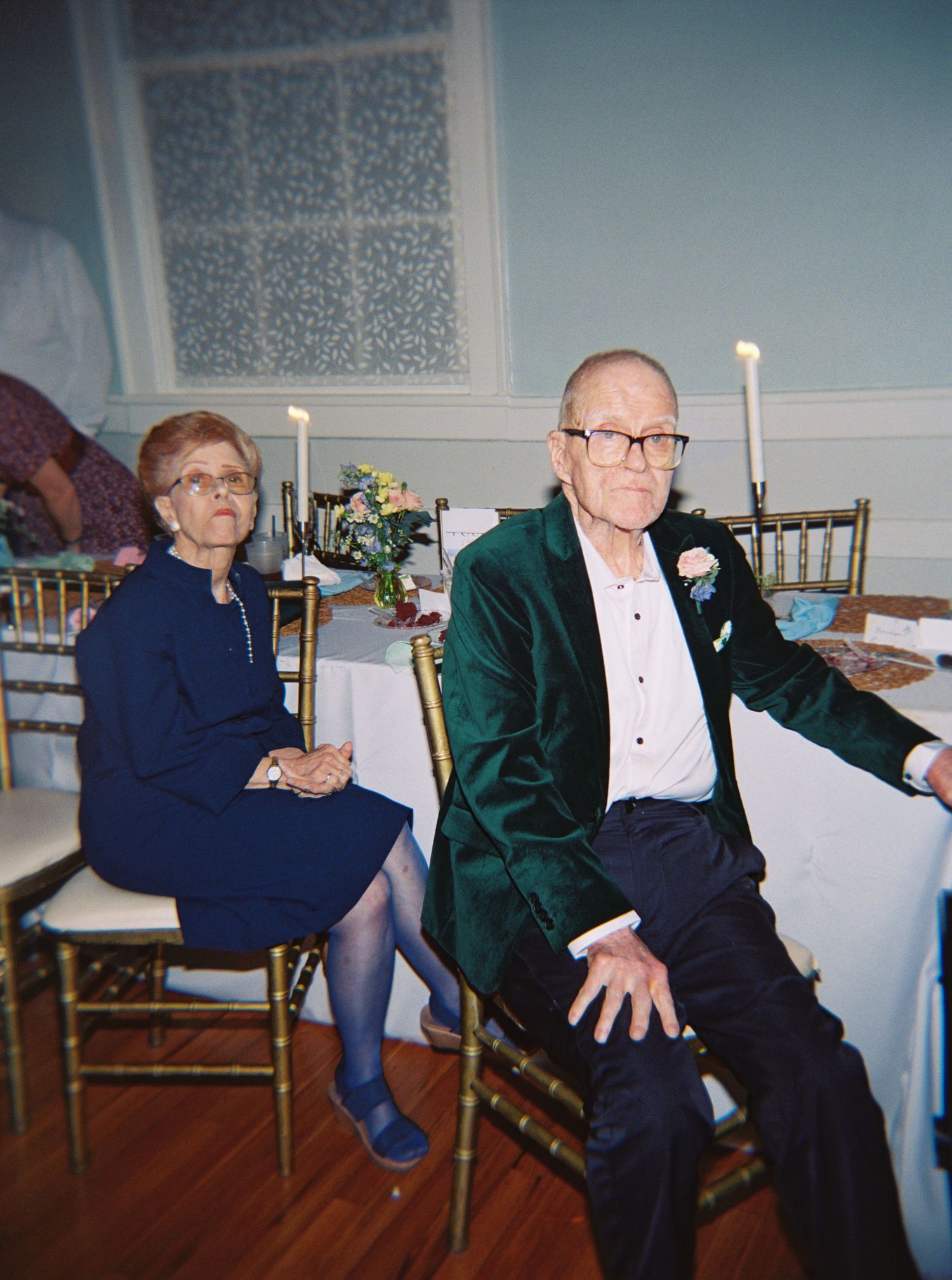 Elderly couple seated at a decorated banquet table, man in green velvet blazer with boutonniere, woman in navy dress.