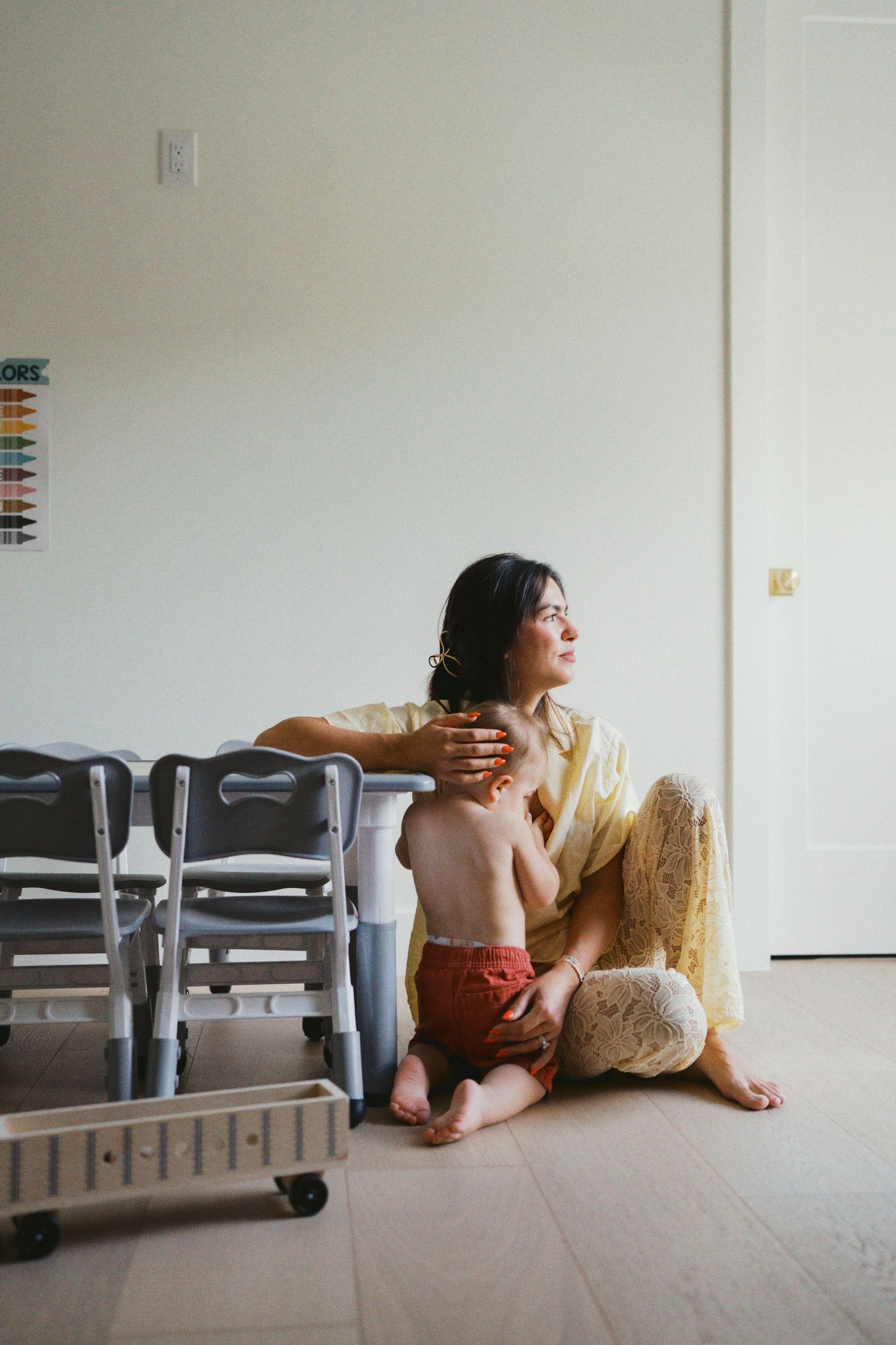 A woman in a yellow dress sits on the floor holding a toddler near small gray chairs in a bright room.