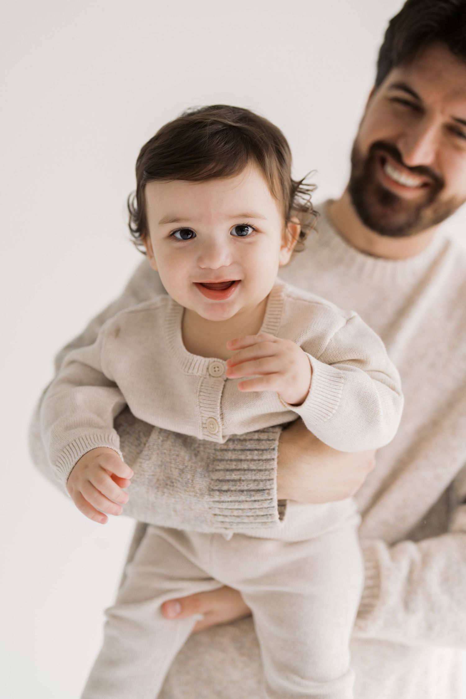 A smiling baby being held by a bearded man in a light-colored sweater against a white background.