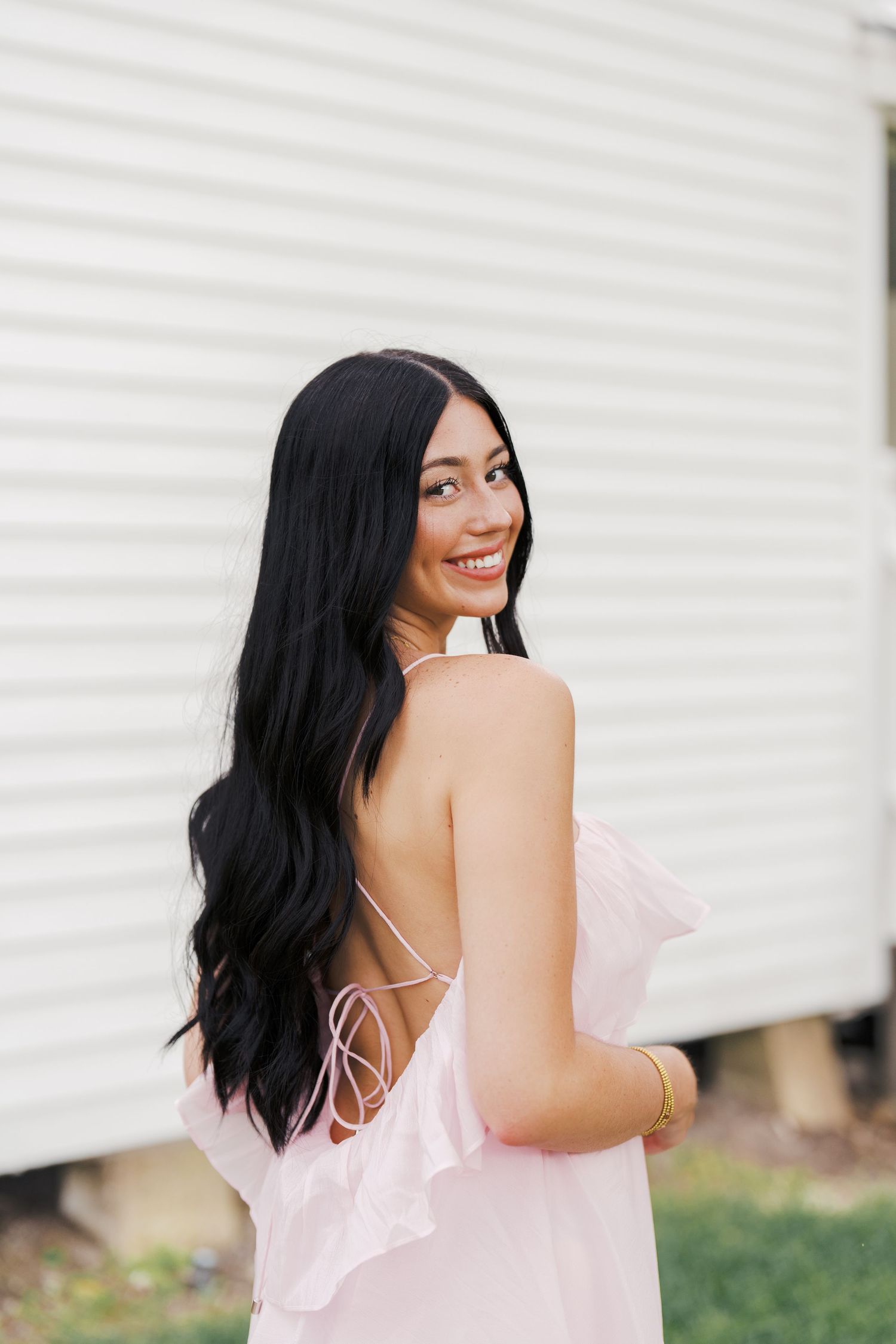 Woman in pink dress with long black hair smiling over her shoulder against white wall background.