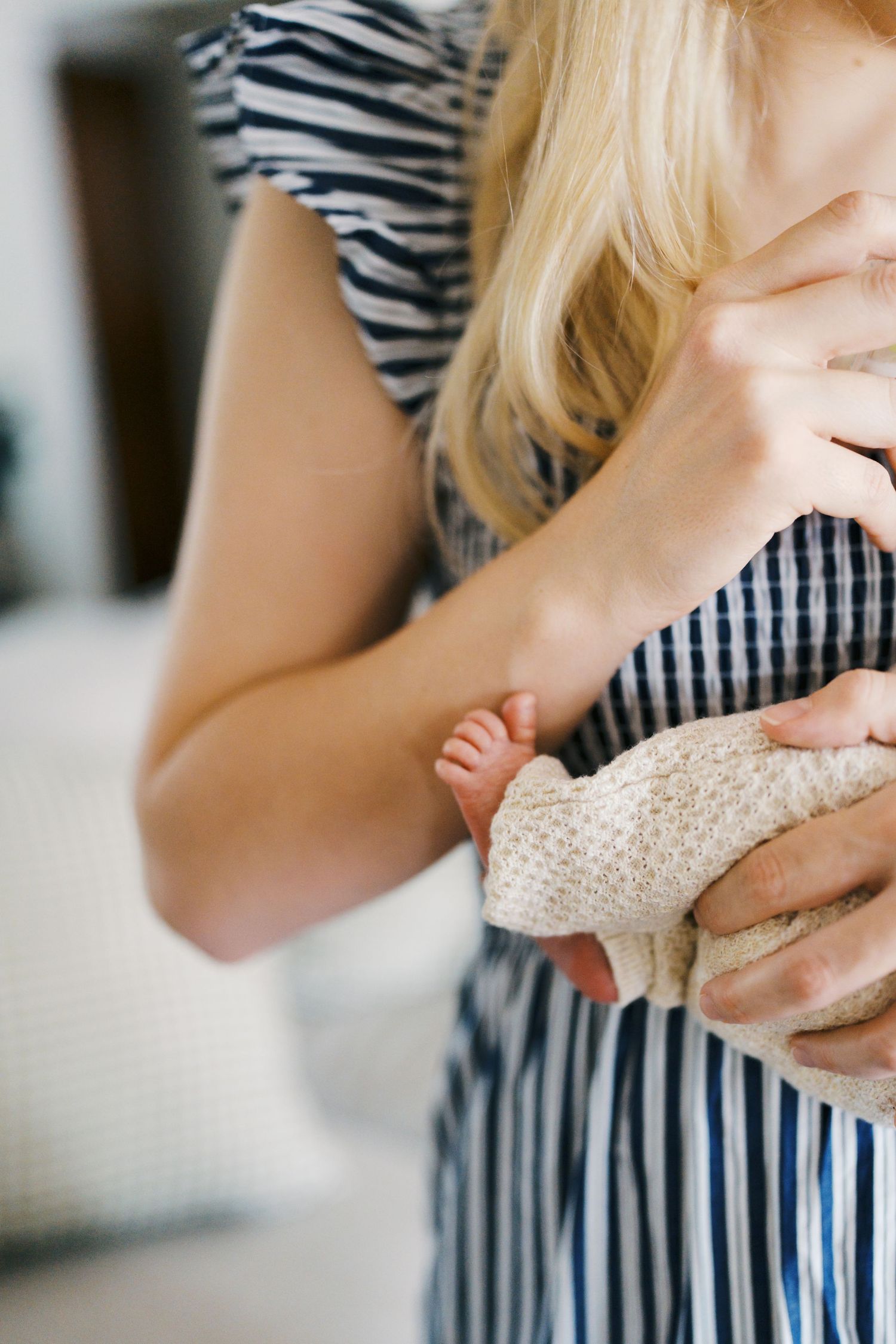 A blonde mother in a striped dress tenderly holds her swaddled newborn baby close to her chest.
