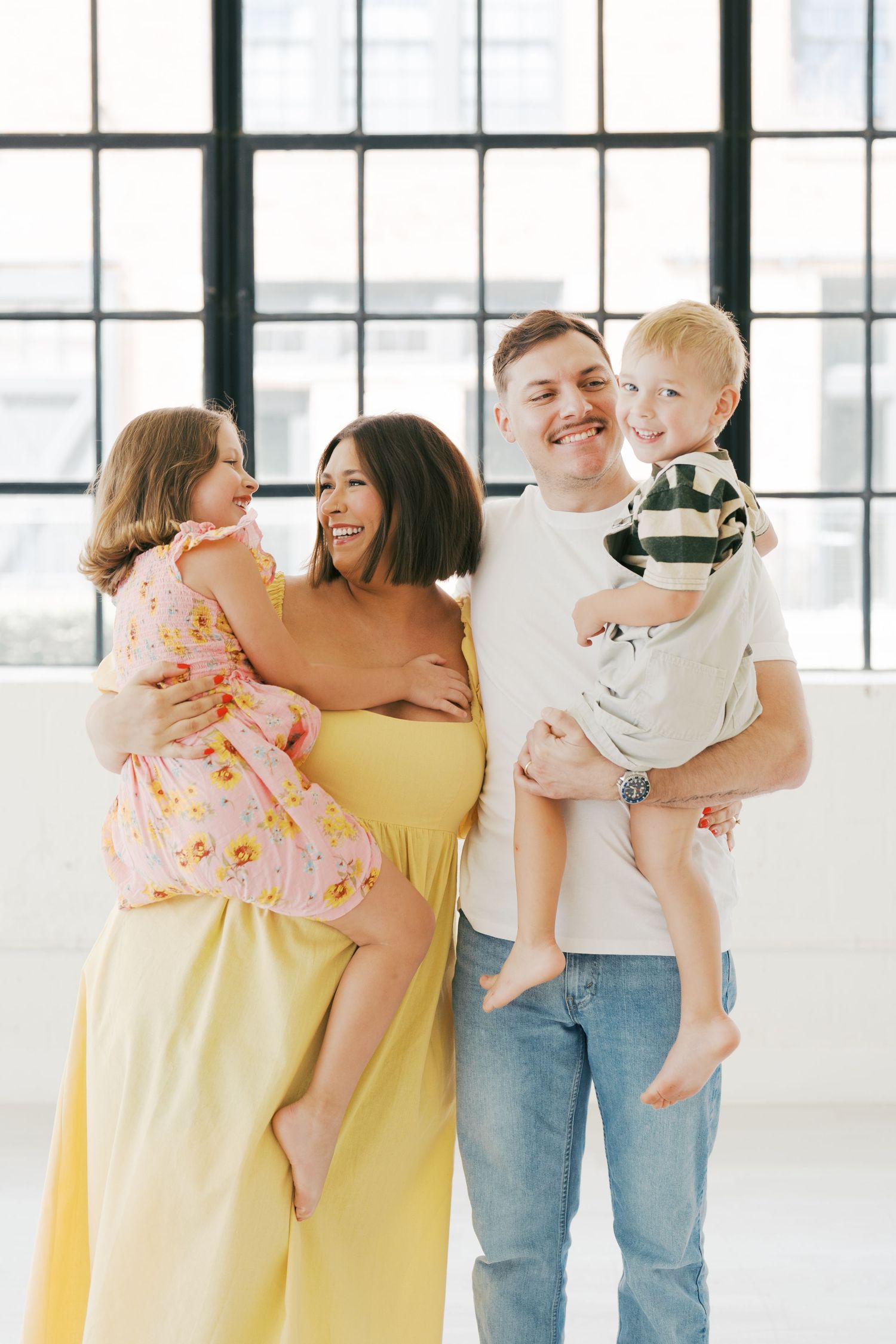 Family gathering shows parents and children in casual outfits of yellow and denim standing together in a bright studio space.
