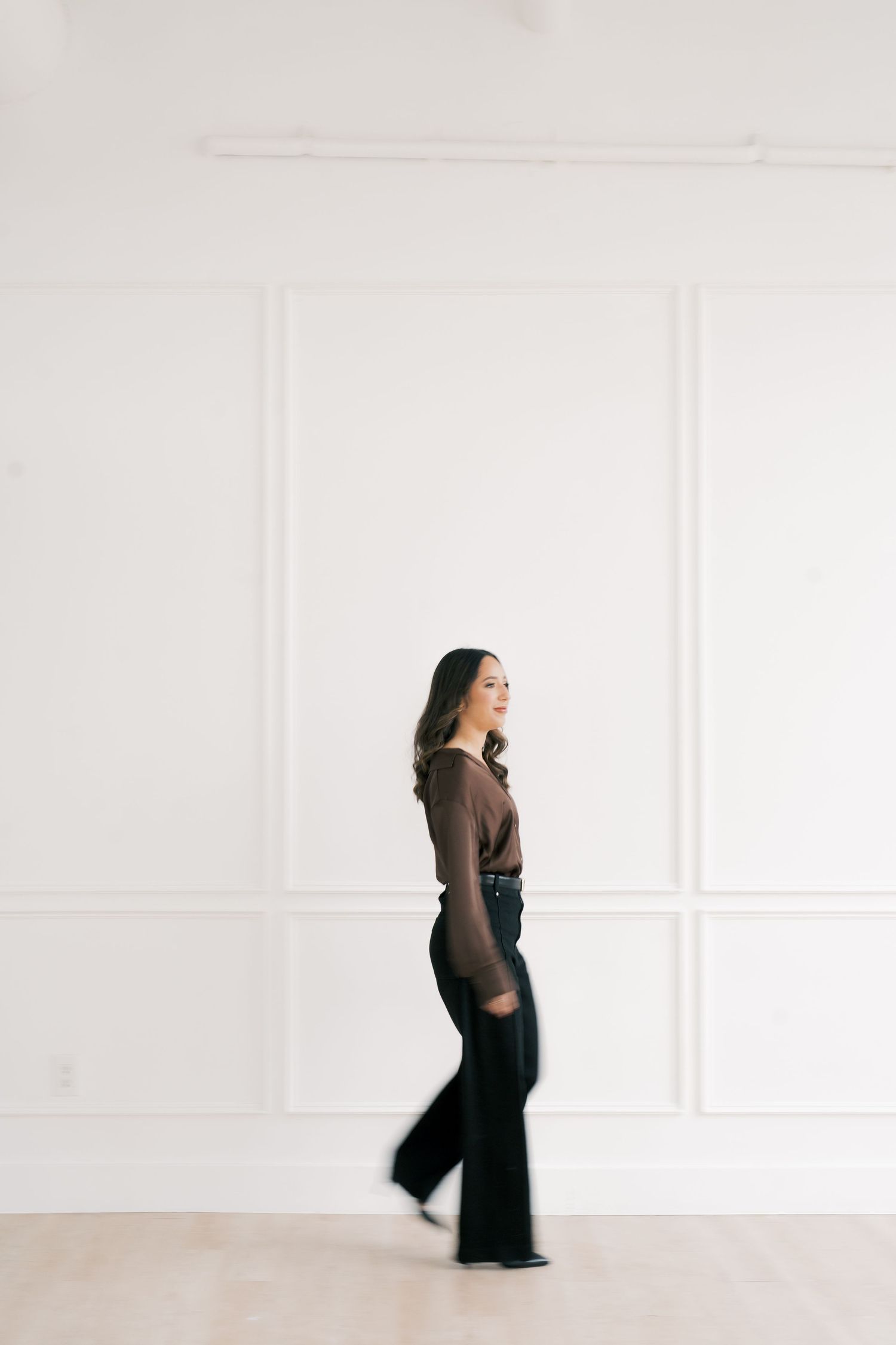 Stylish figure in brown top and black pants poses against white paneled wall in minimal setting.