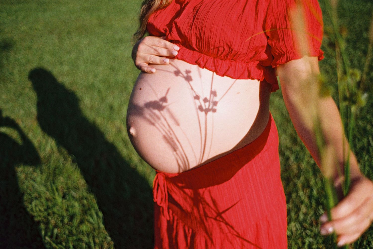 Pregnant woman in red dress outdoors, cradling bare baby bump with wildflower shadows on skin in sunny green field.
