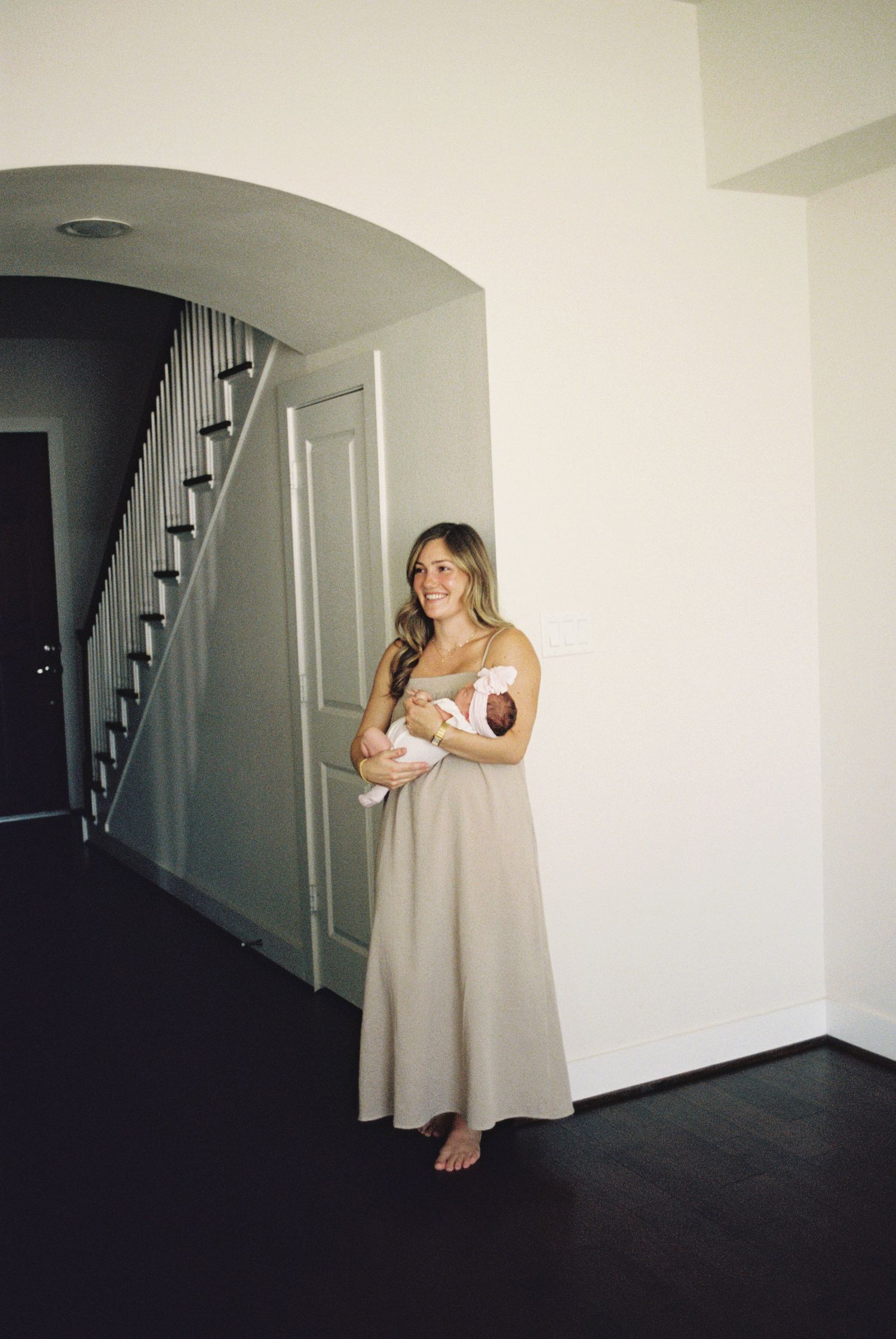 Woman in beige maxi dress holding newborn baby in bright home hallway with staircase.
