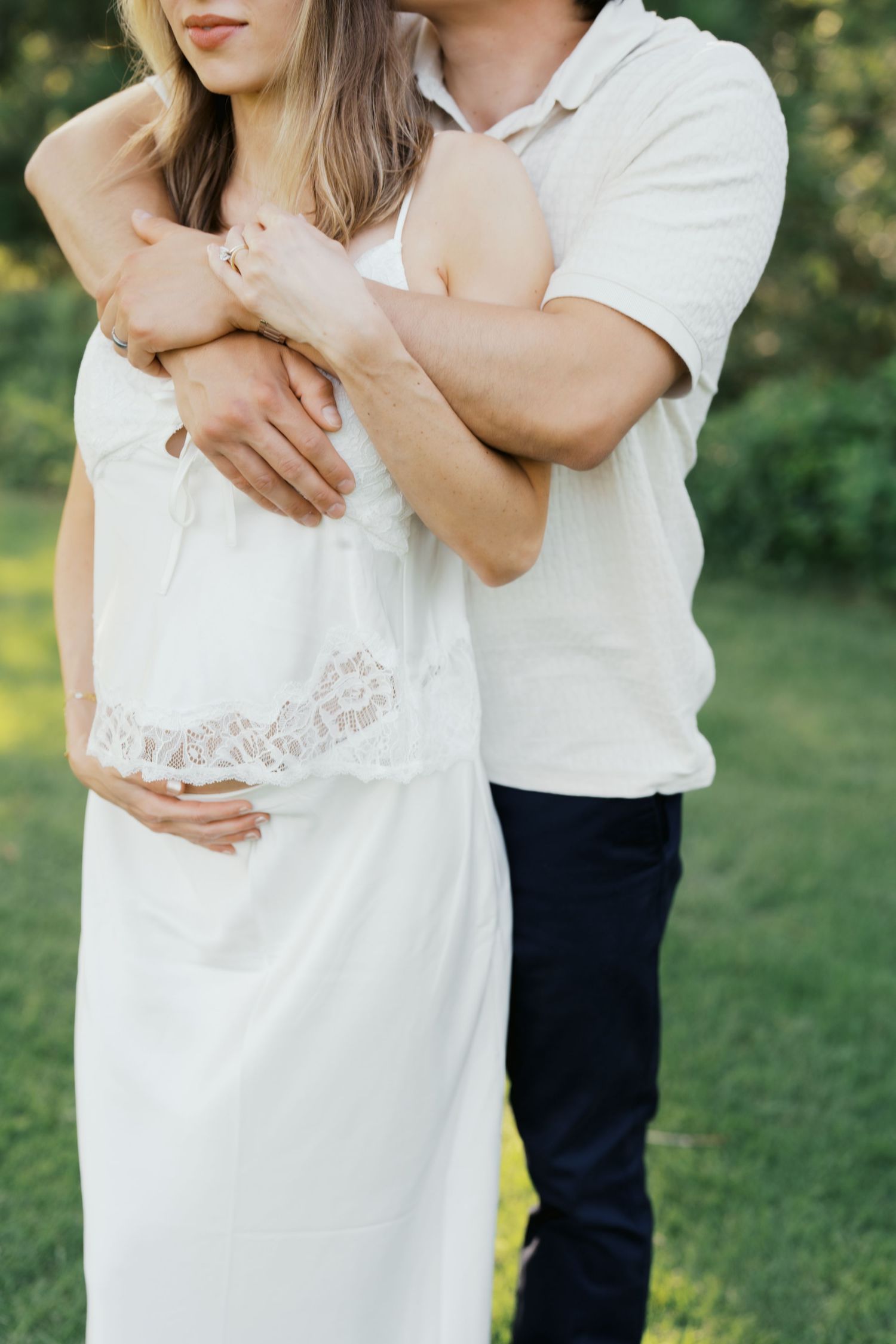 A couple embracing outdoors, pregnant woman in white lace dress with partner's arms wrapped around her baby bump.