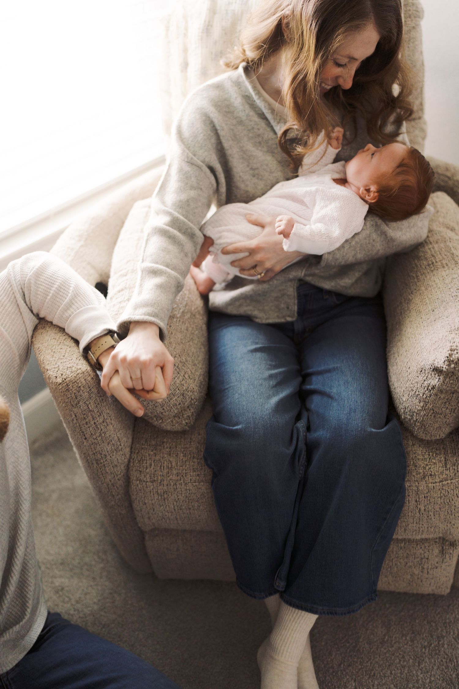 A mother sits in an armchair cradling her newborn baby, wearing jeans and a gray sweater near a bright window.