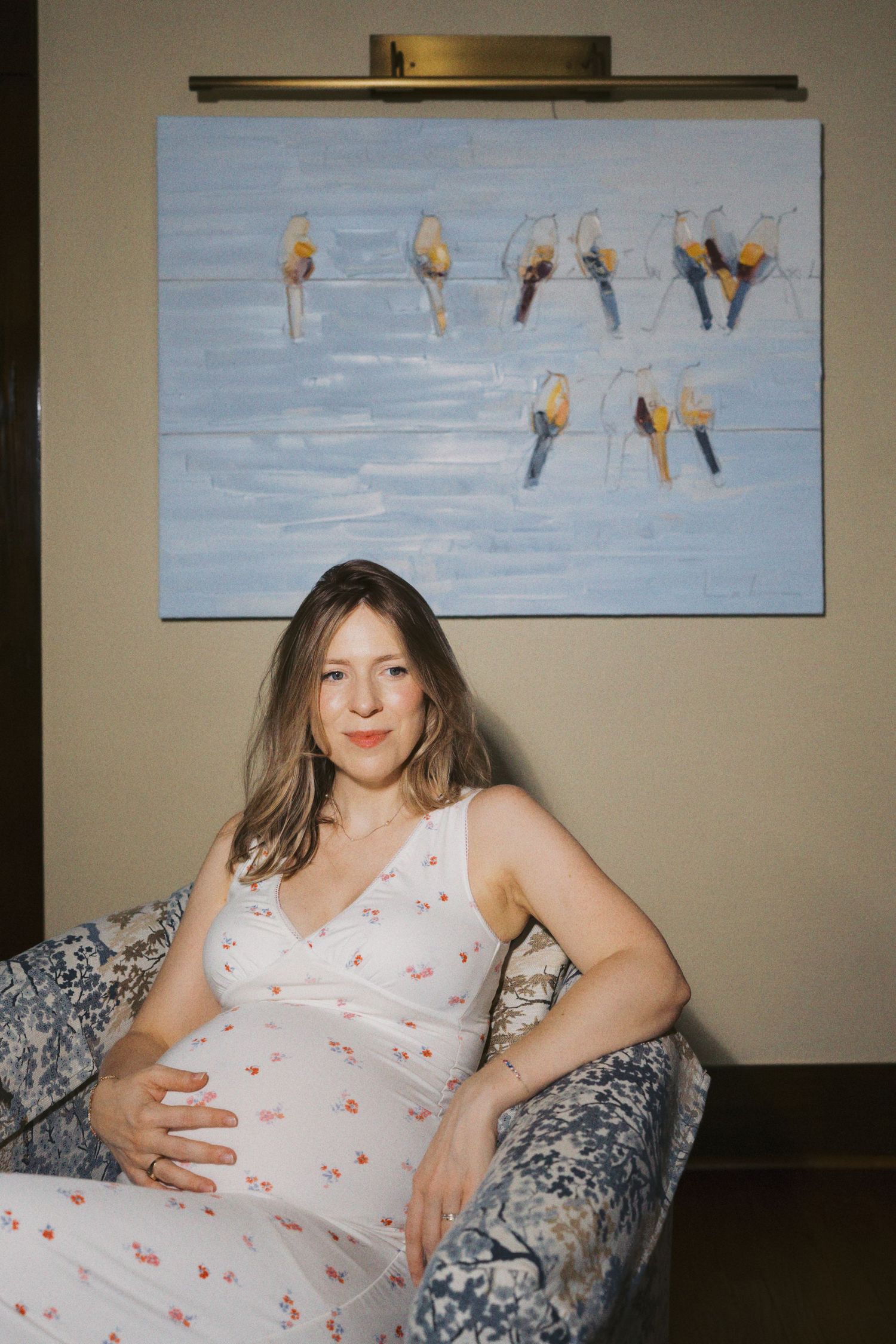 Pregnant woman in floral dress sitting in blue patterned chair with bird artwork on wall behind her.