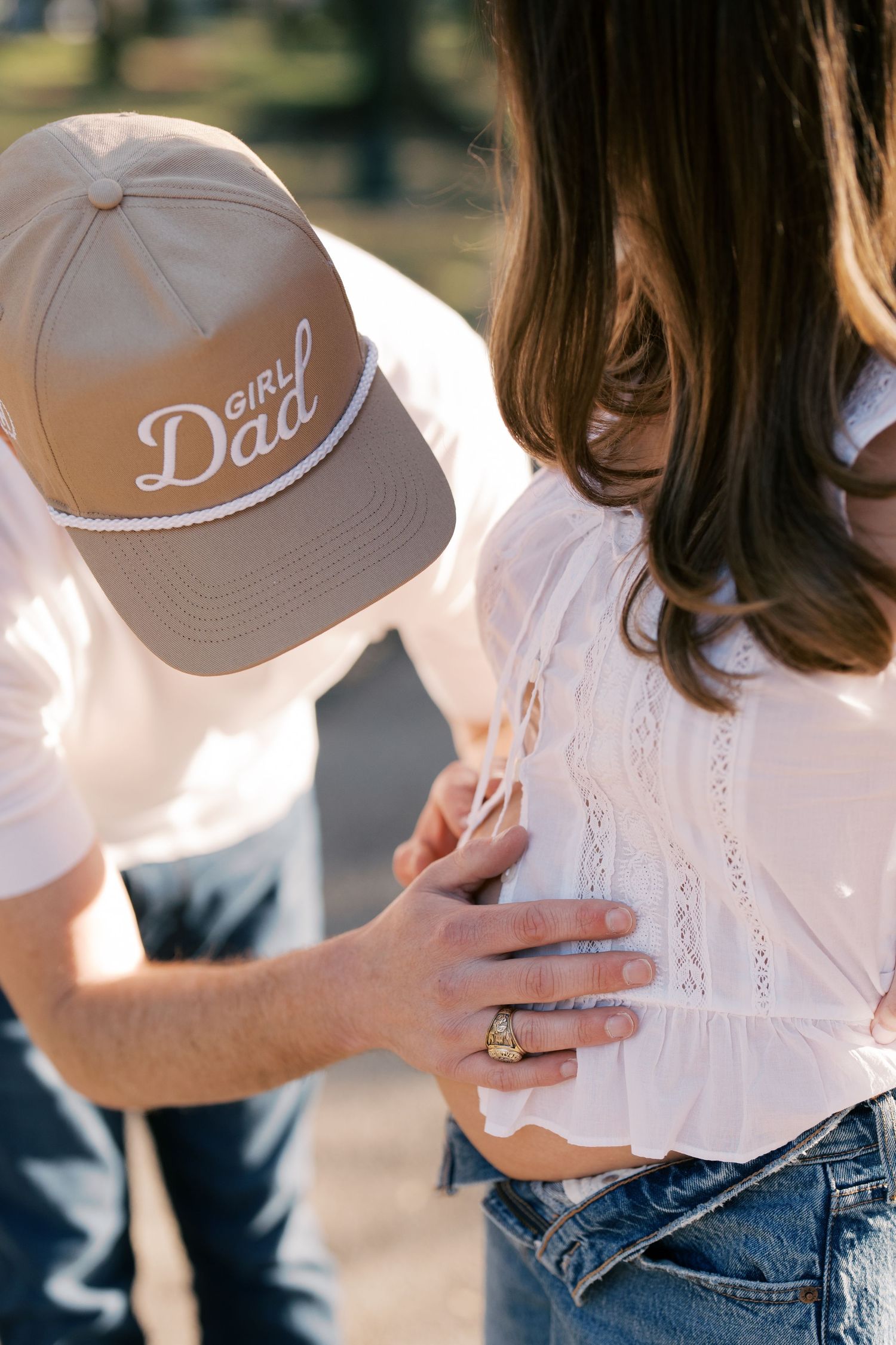 A young woman wearing a white cap with the text 'Daddy's Girl' is standing with a man in an outdoor setting.