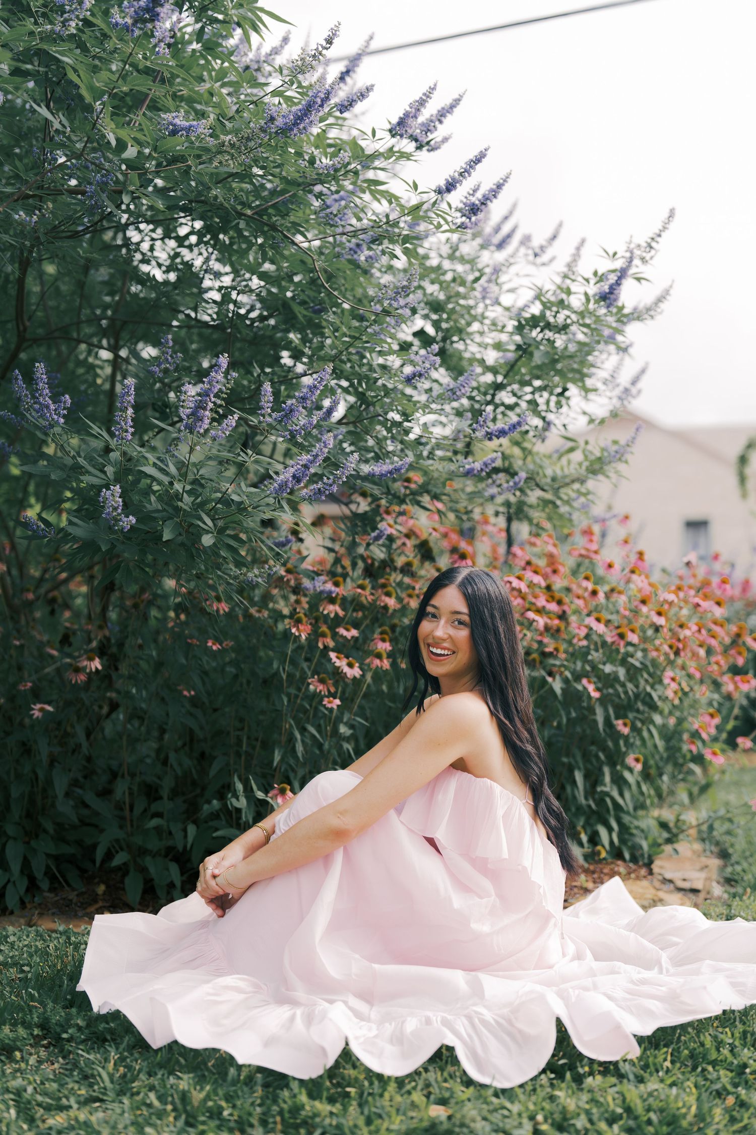 Model in flowing pink dress sitting in garden with purple flowers and greenery.