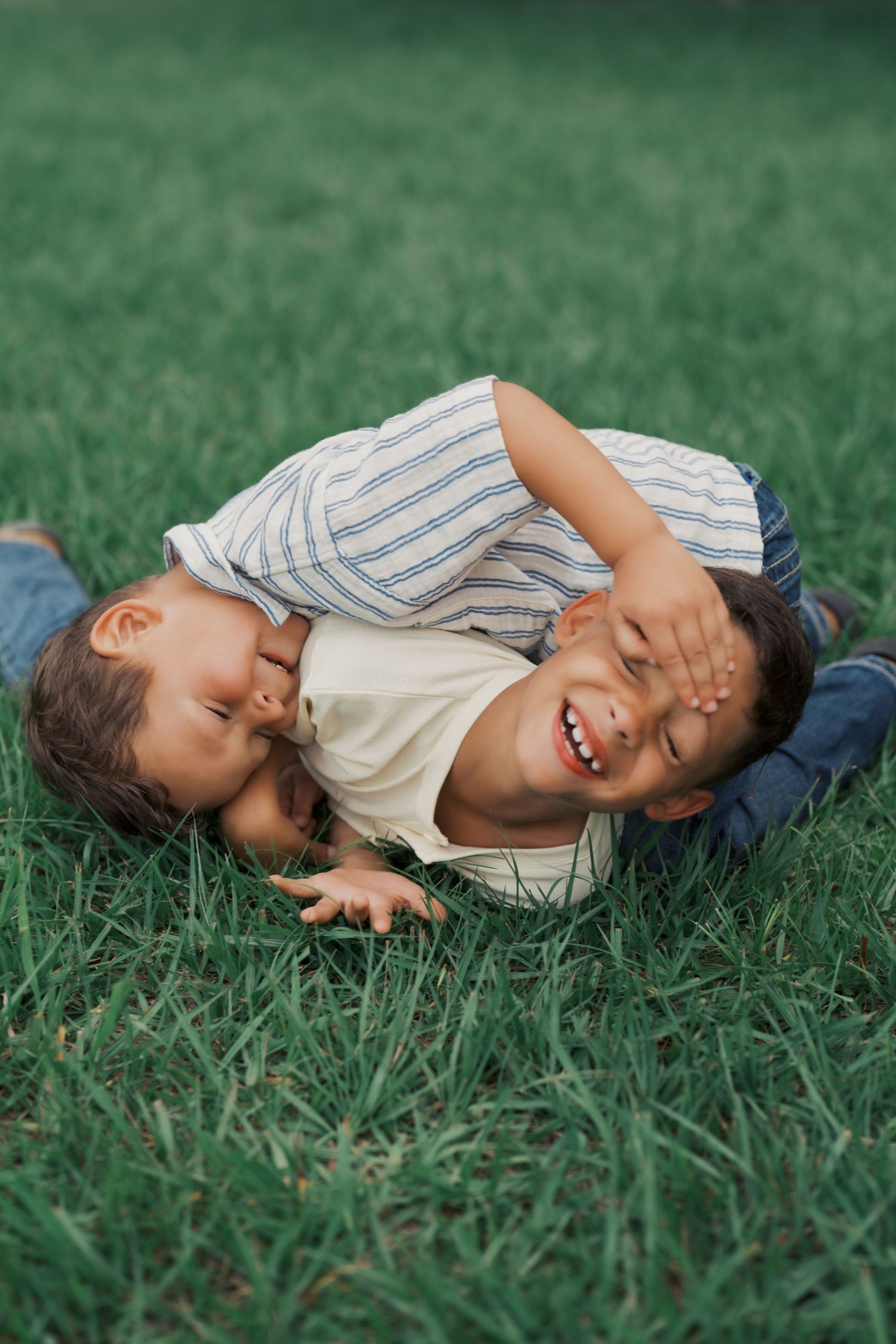 Two young boys laughing and playing together on green grass, enjoying a fun outdoor wrestling match.