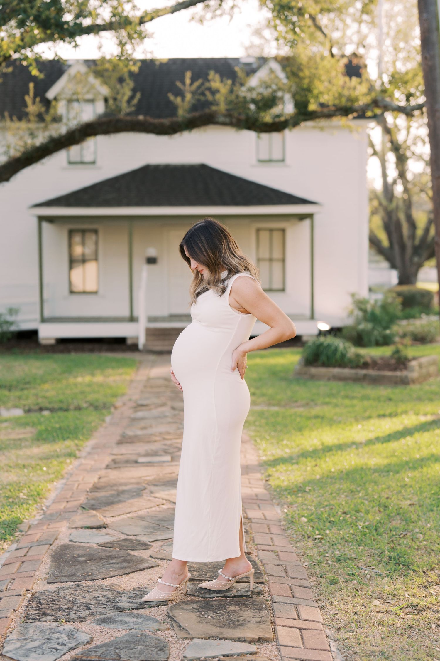 Expectant mother in a flowing white dress stands on a stone path in front of a charming white farmhouse at sunset.