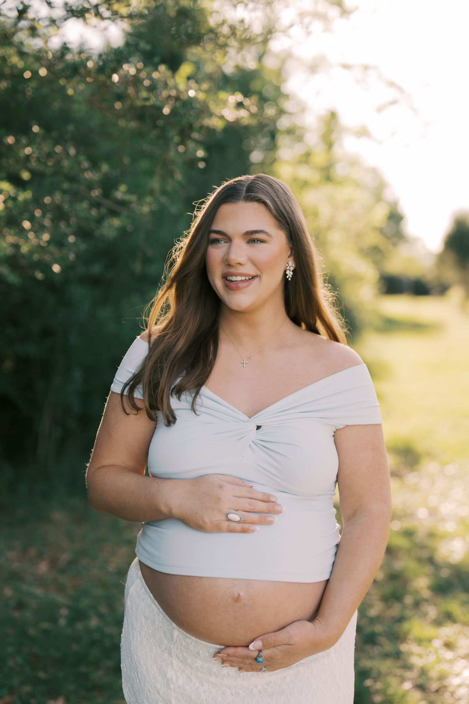 Smiling pregnant woman in white off-shoulder crop top cradling baby bump outdoors in golden sunlight.