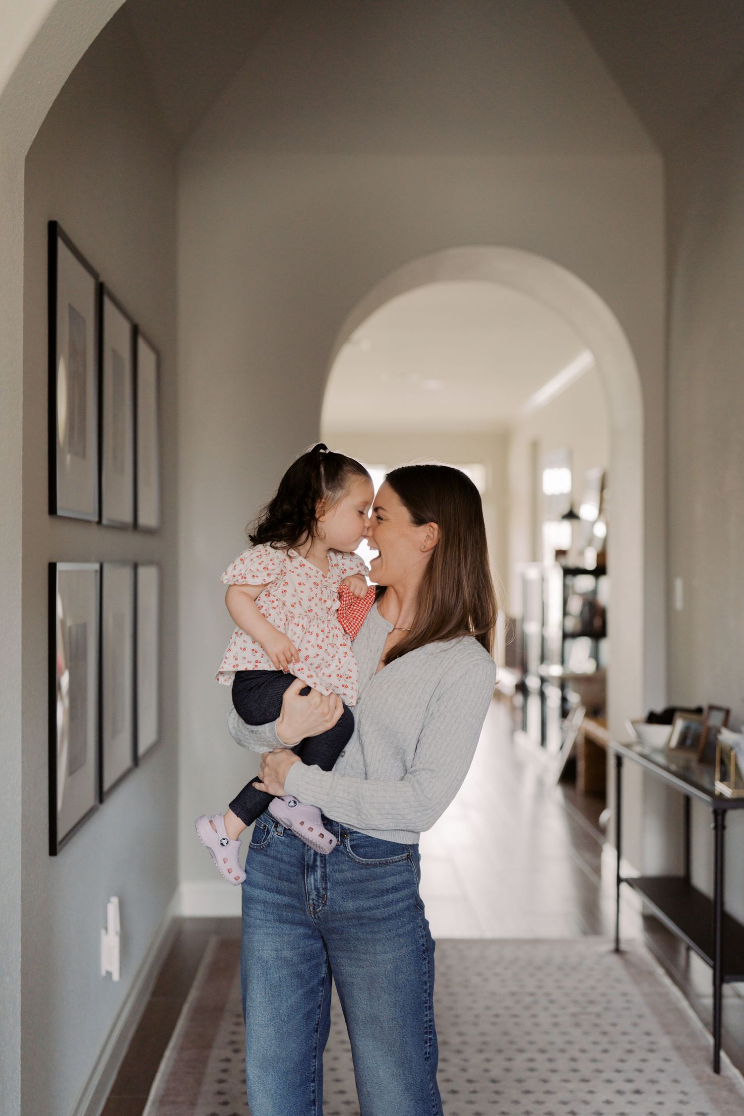 A woman in a gray sweater holds and embraces a young girl in a hallway with framed artwork on the walls.