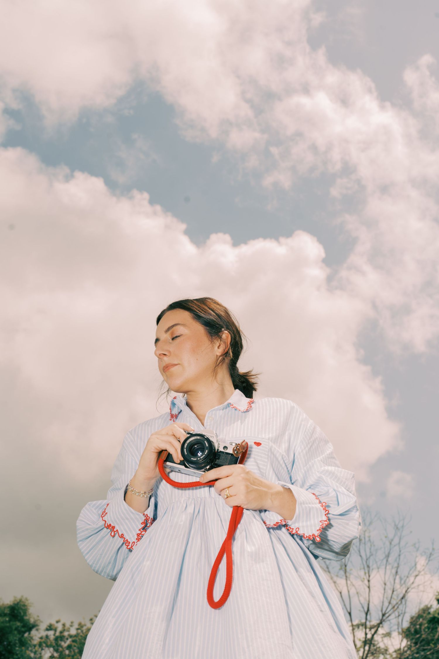 Woman in striped dress holding vintage camera with red strap against a cloudy sky backdrop.