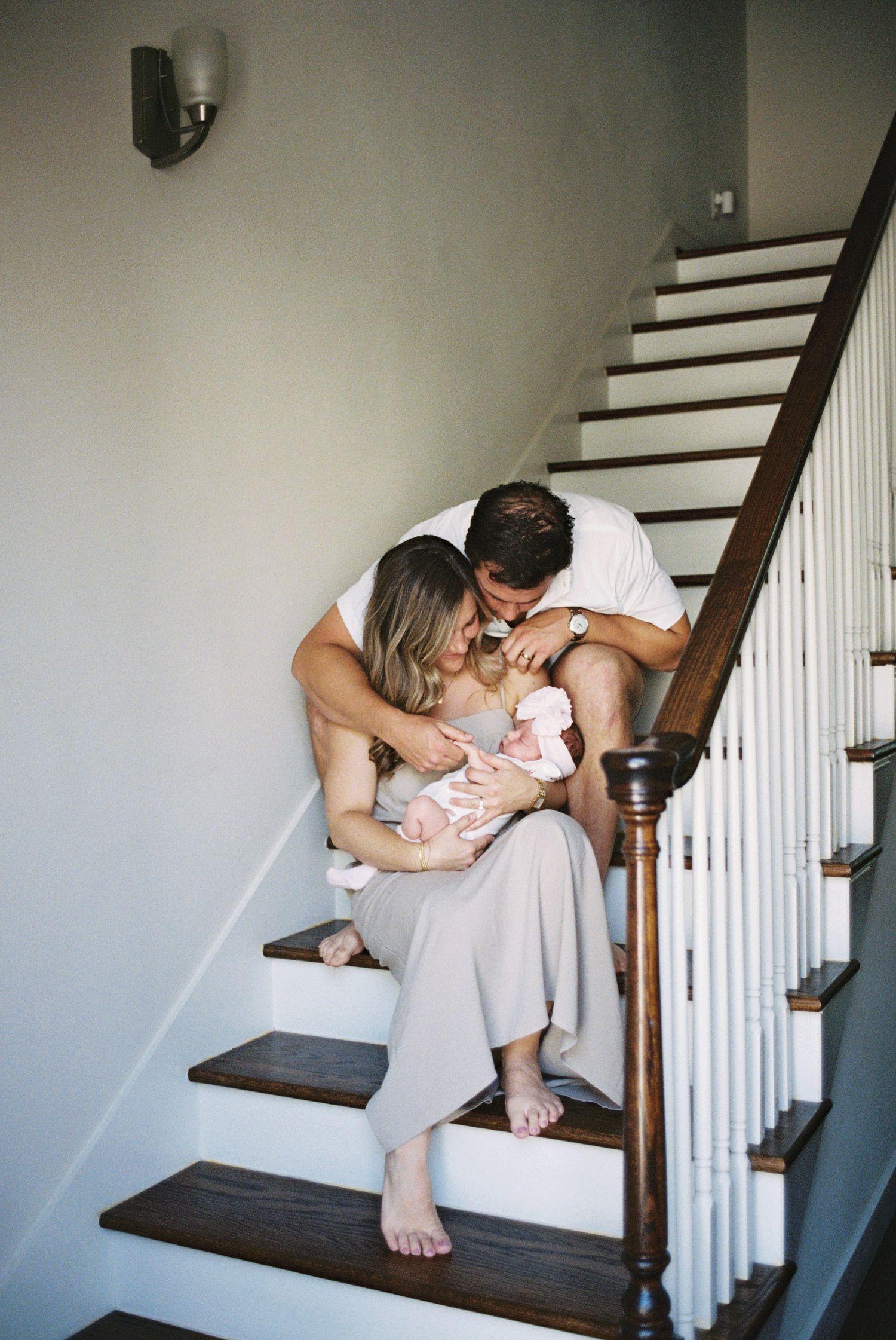 A couple sits on wooden stairs, lovingly gazing at their newborn baby wrapped in pink.