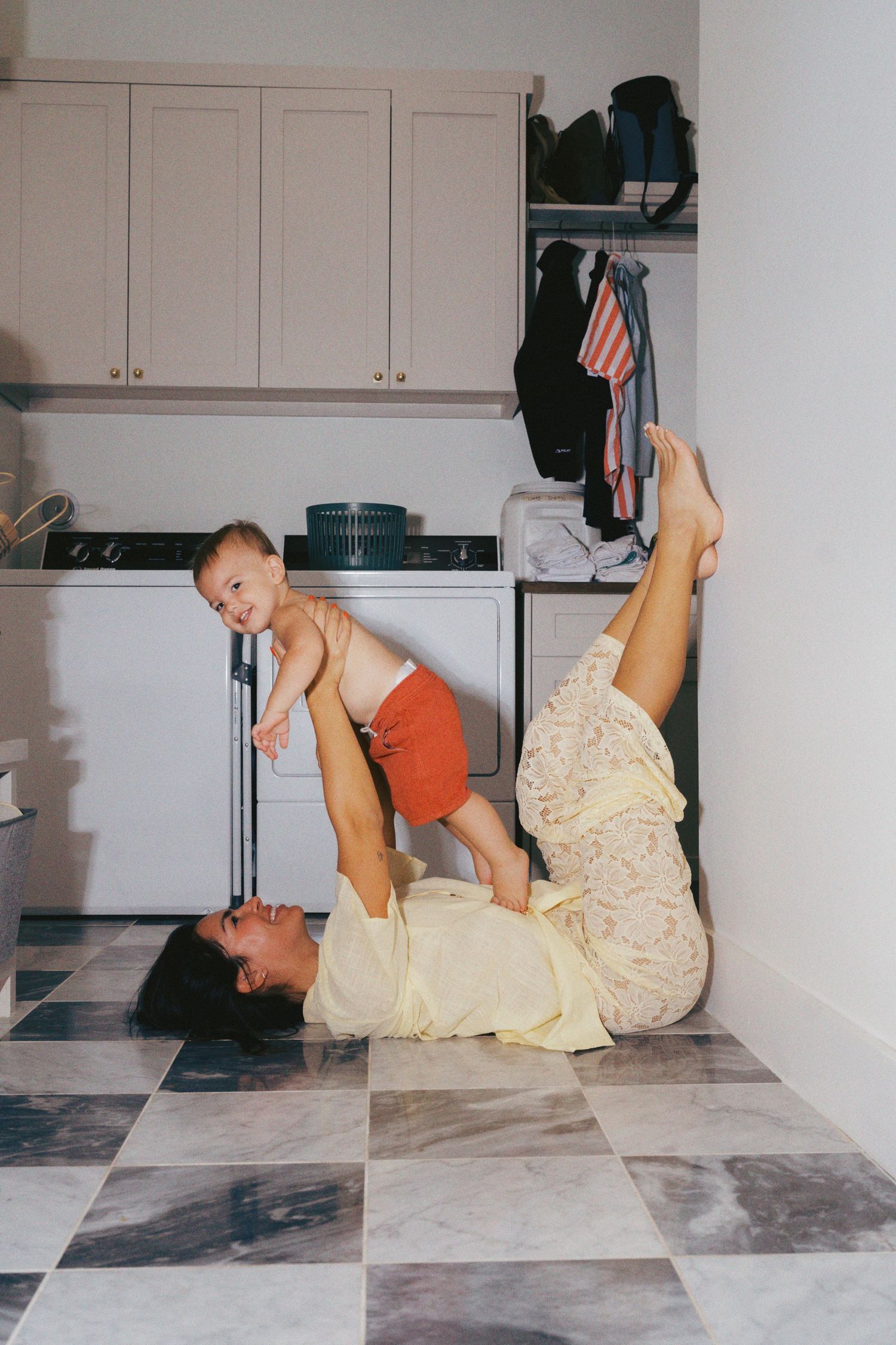 Mother lying on kitchen floor playfully lifting toddler in red shorts above her with her feet in a joyful moment.