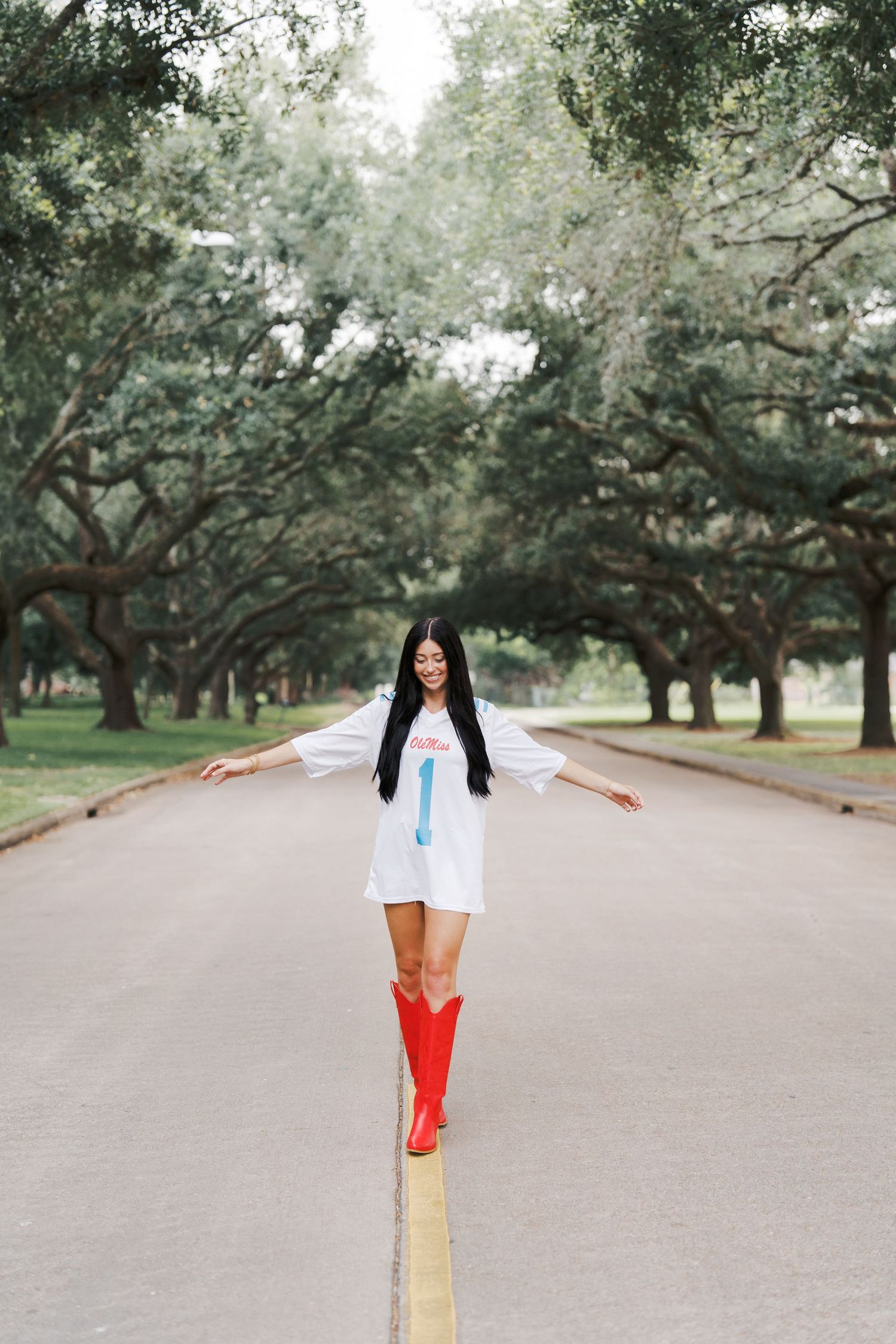 Person in white dress and red boots balancing on yellow line under oak trees.
