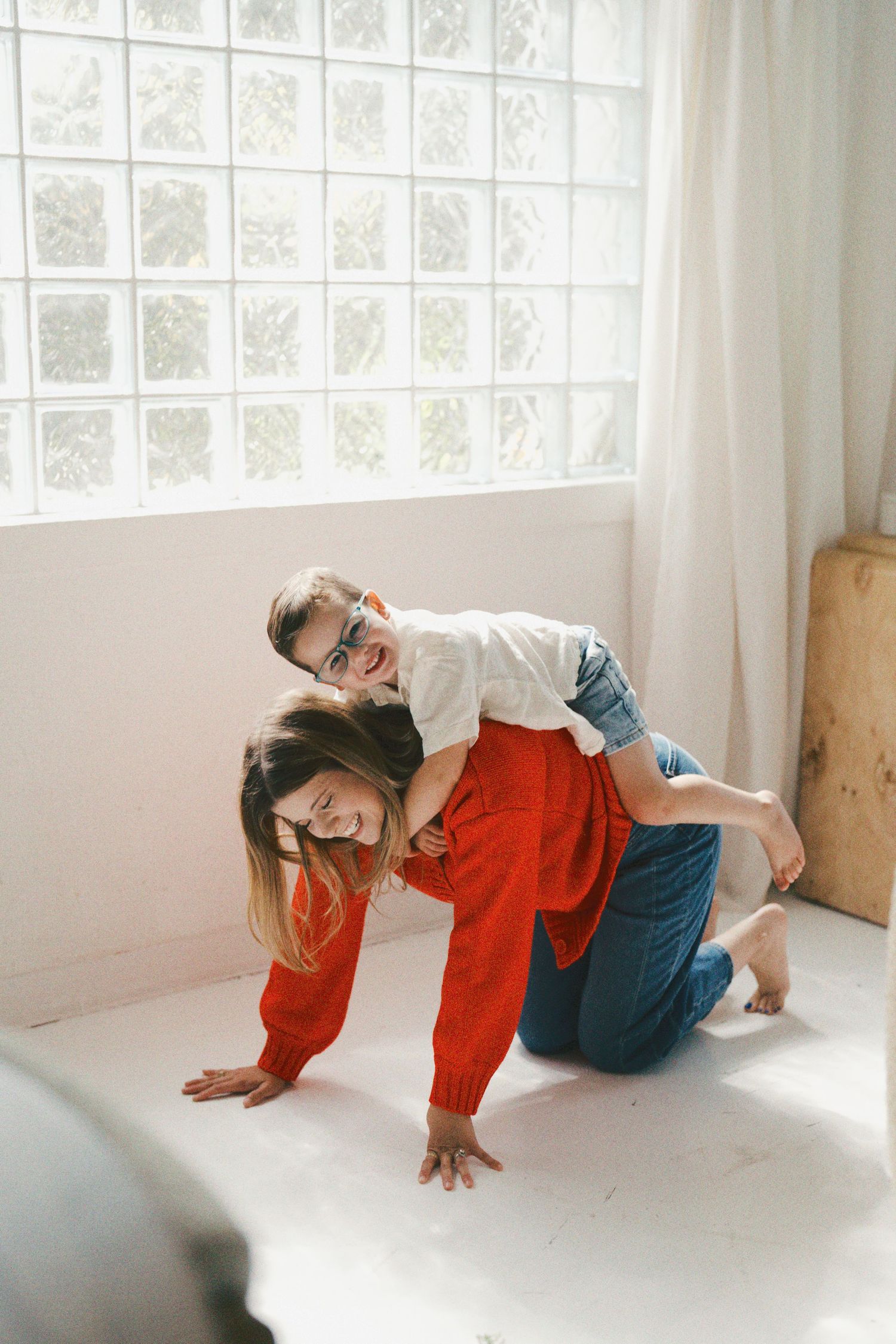 A woman in a red sweater plays with a child on her back, both laughing in a bright white room.
