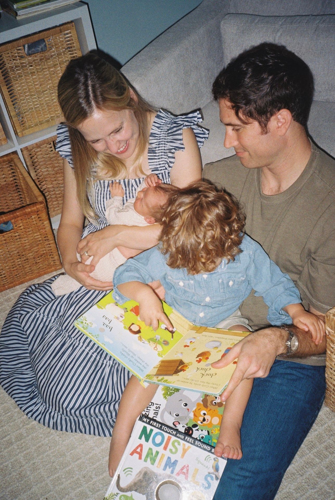 Family reading Noisy Animals book together, parents sitting on floor with two young children in cozy home setting.