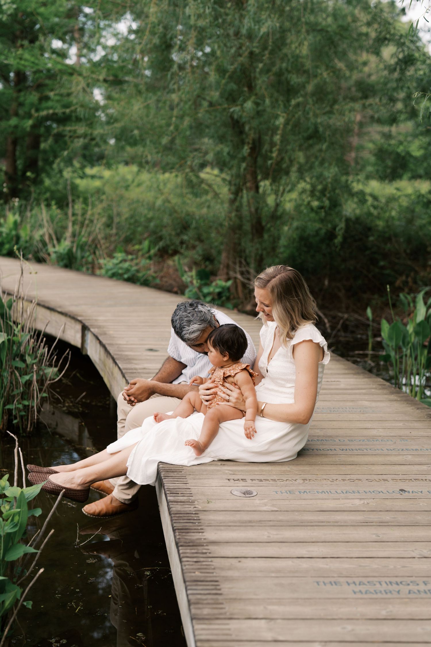 A family of three sits on a wooden bridge surrounded by lush greenery, parents lovingly holding their baby.