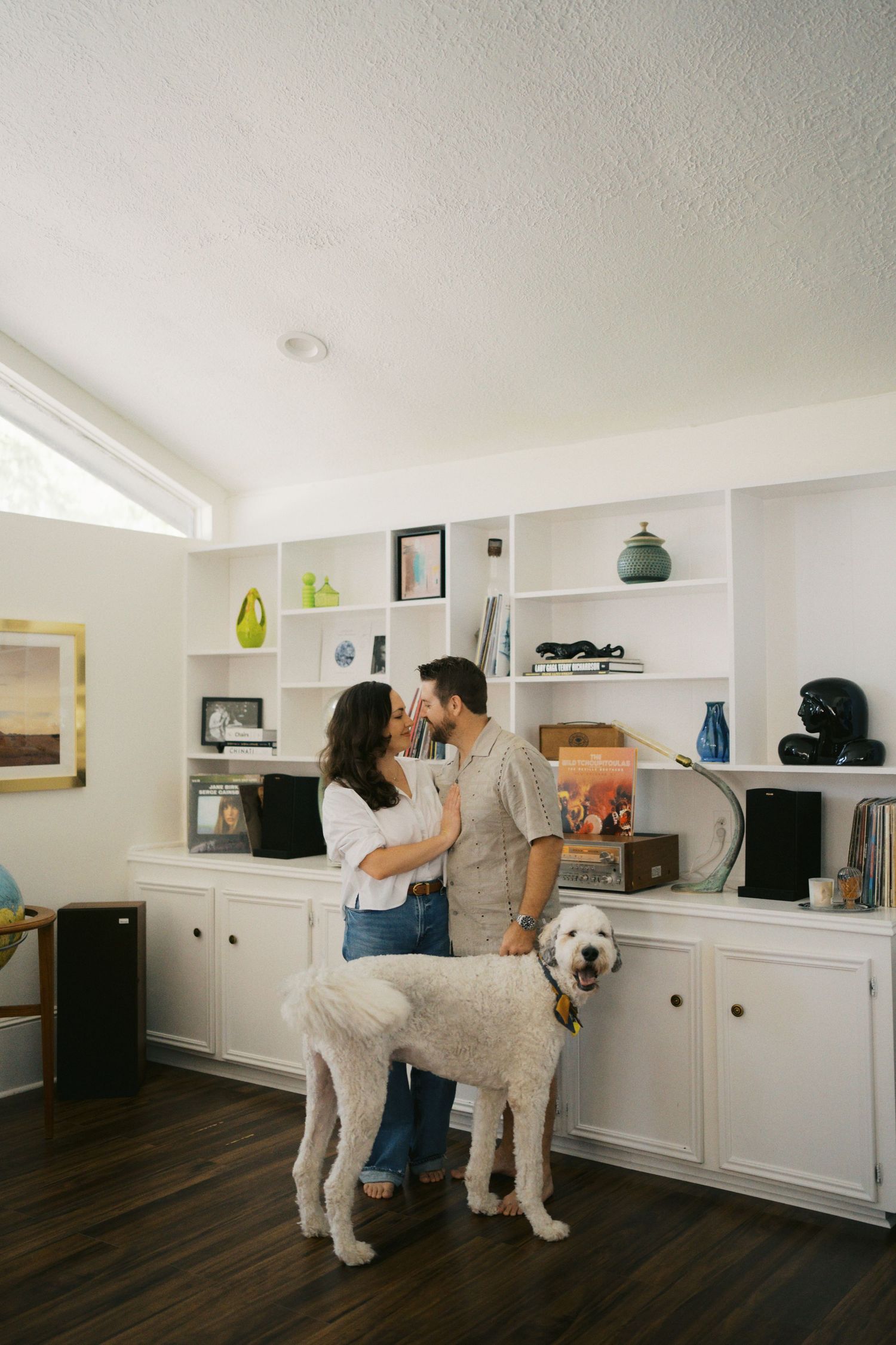 Couple sharing a romantic moment in a modern home office with built-in shelves and a large white dog standing beside them.