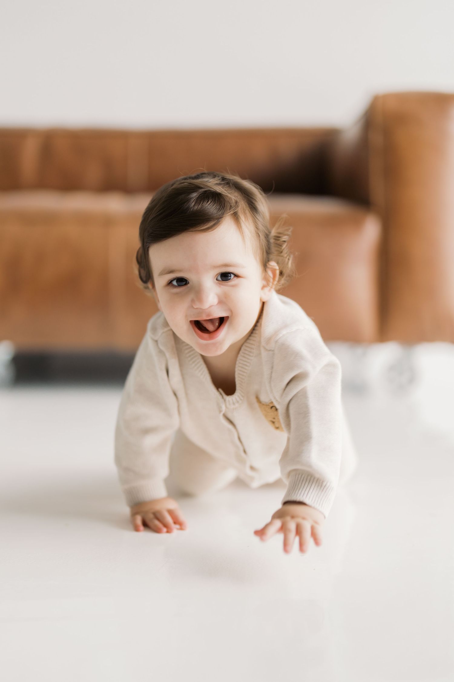A smiling baby wearing a white sweater crawls on a white floor with a wooden couch in the background.