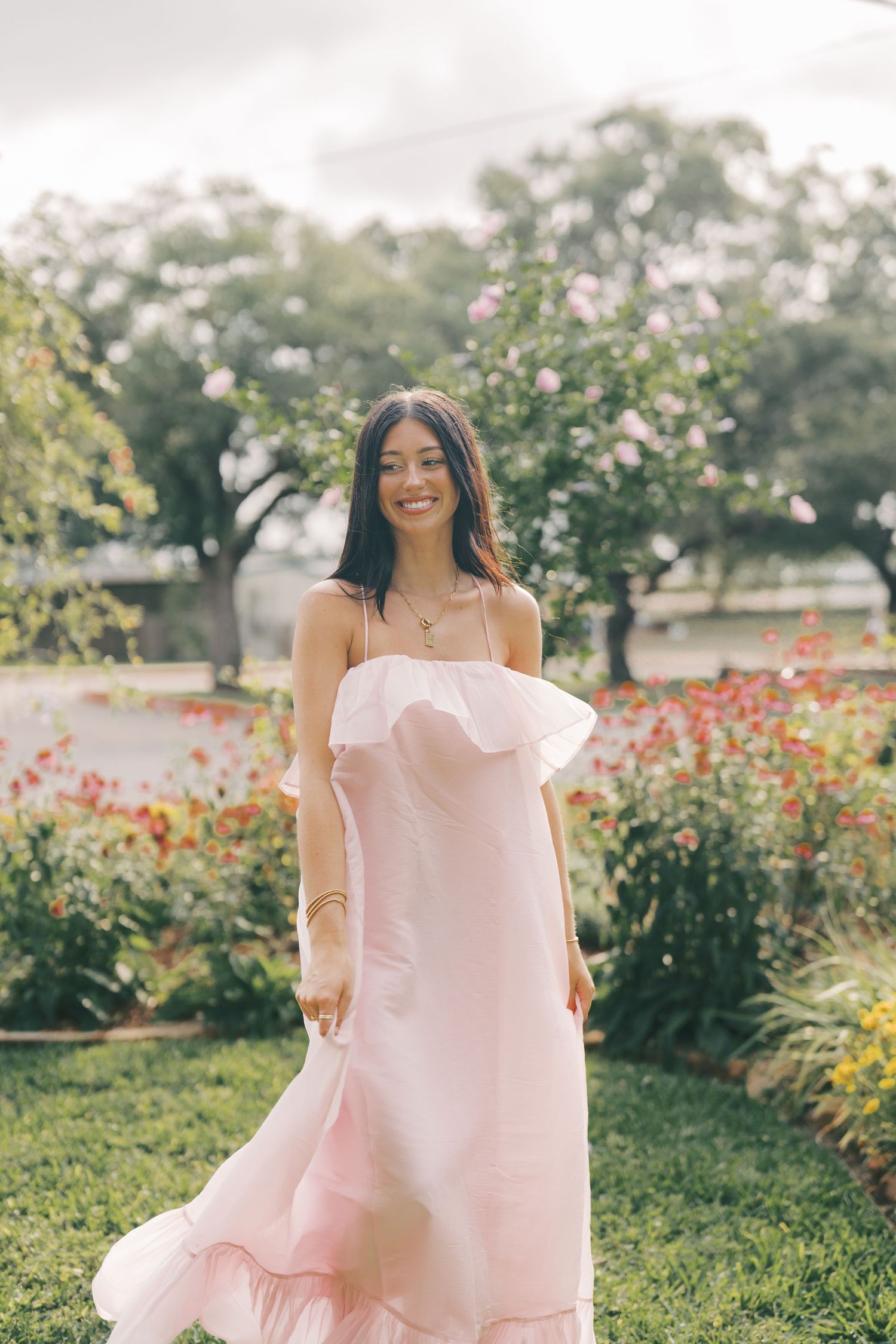 Woman in light pink maxi dress standing in blooming garden with red flowers.