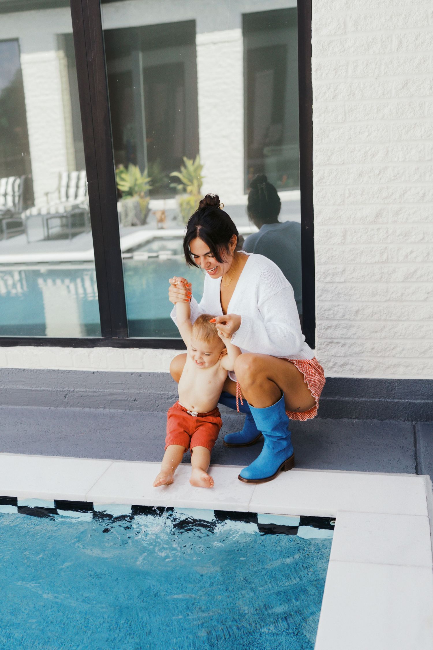 Mother helps toddler splash feet in pool while wearing blue rain boots outdoors.