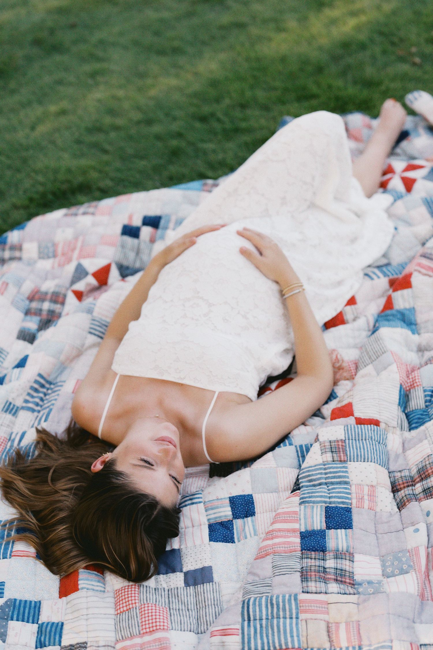 Woman in white lace dress lying on a patchwork quilt outdoors on green grass.