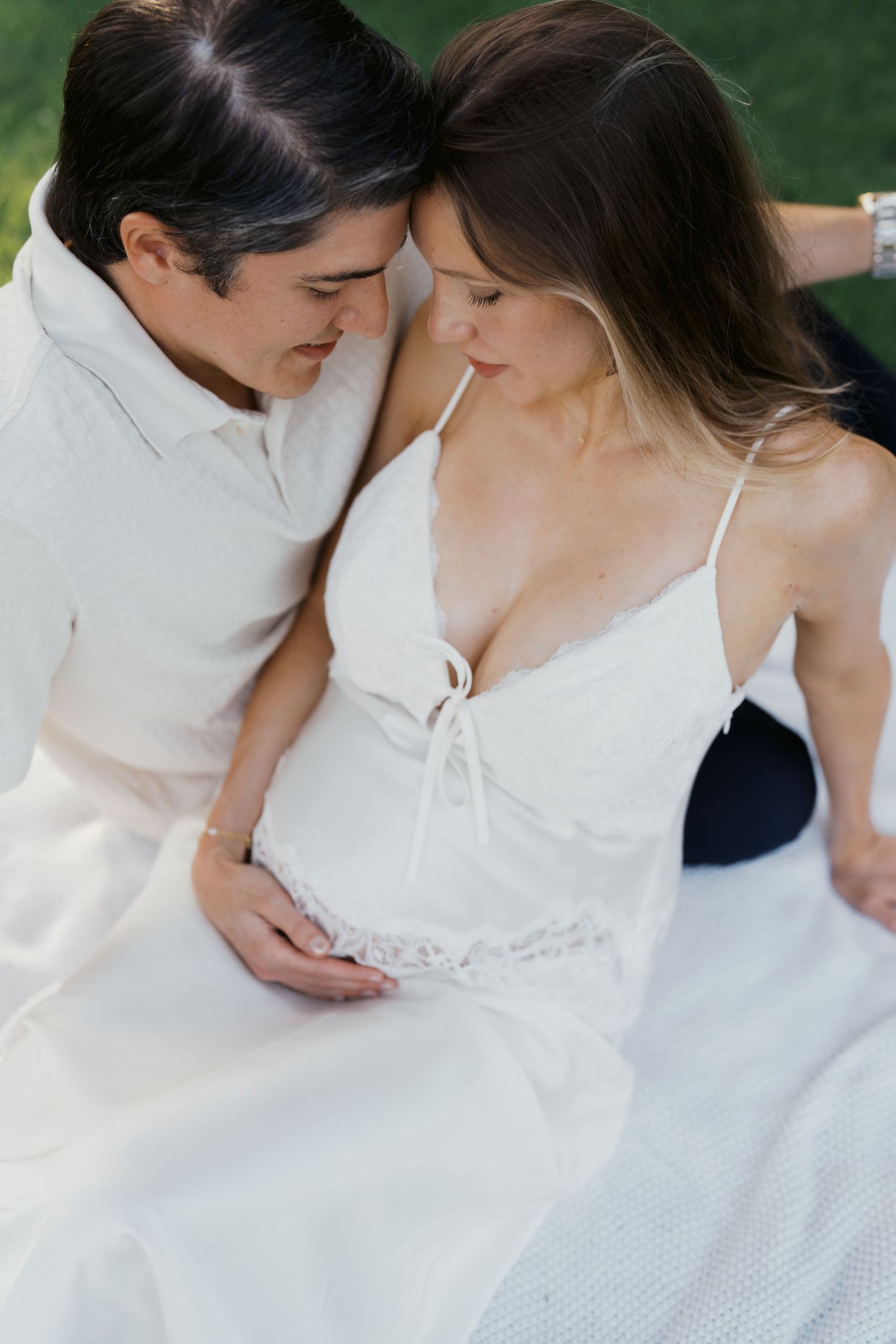 Couple sharing a tender moment outdoors, woman in white lace maternity dress, man in white shirt cradling her baby bump.