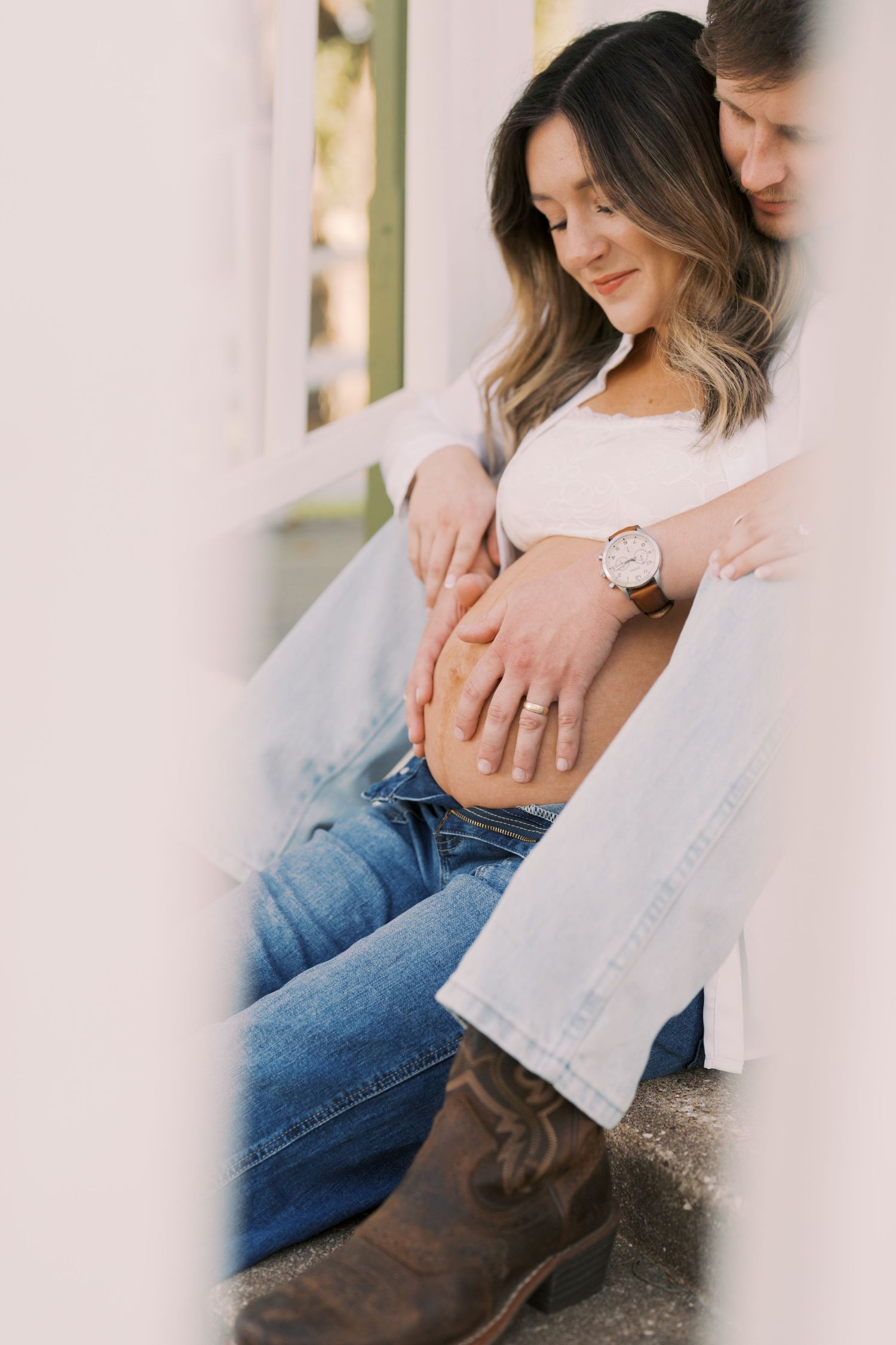 Romantic couple sitting together in casual denim and white outfits showing engagement ring in natural lighting.