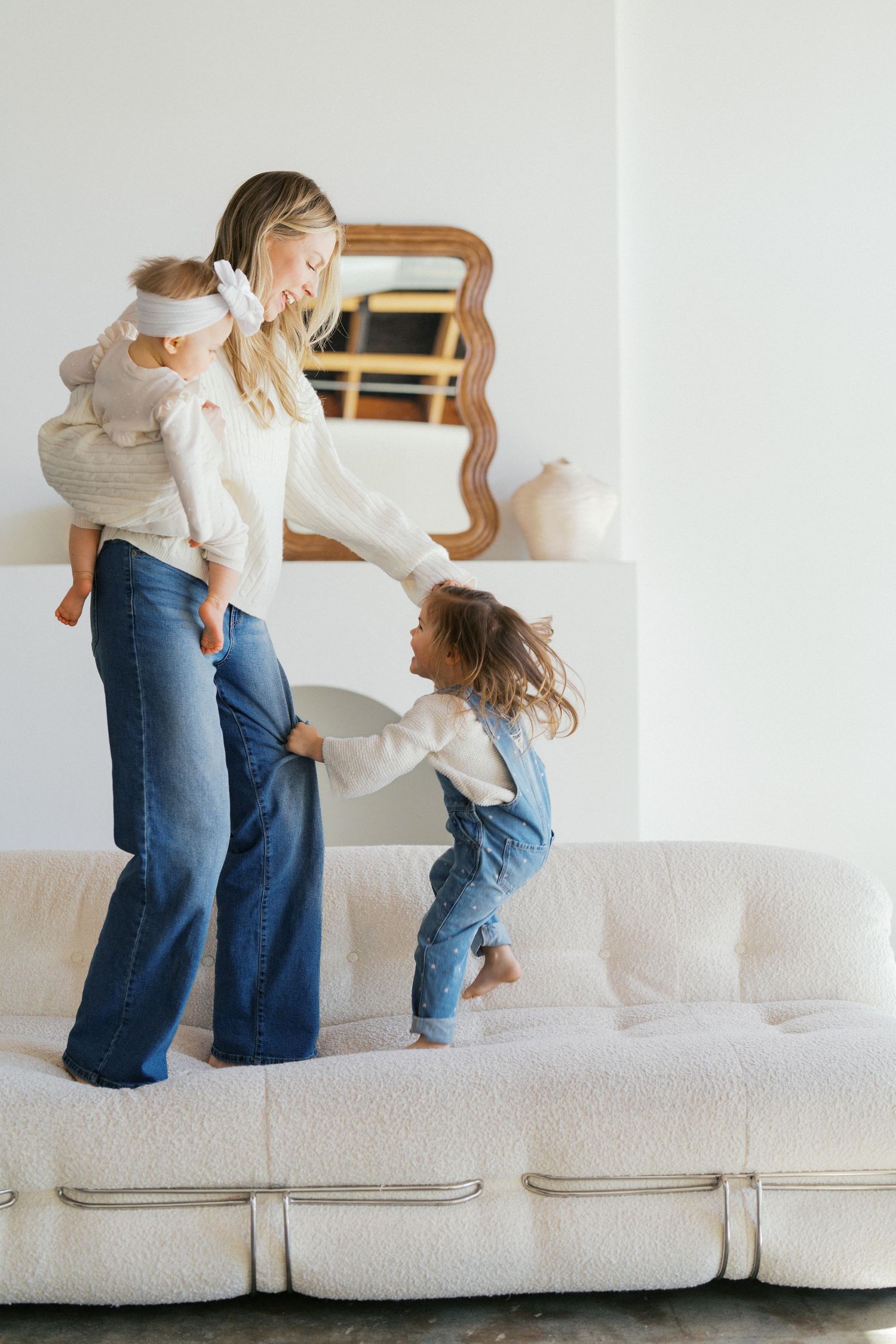 Two people in casual clothing playing and jumping together on a plush white sectional sofa.