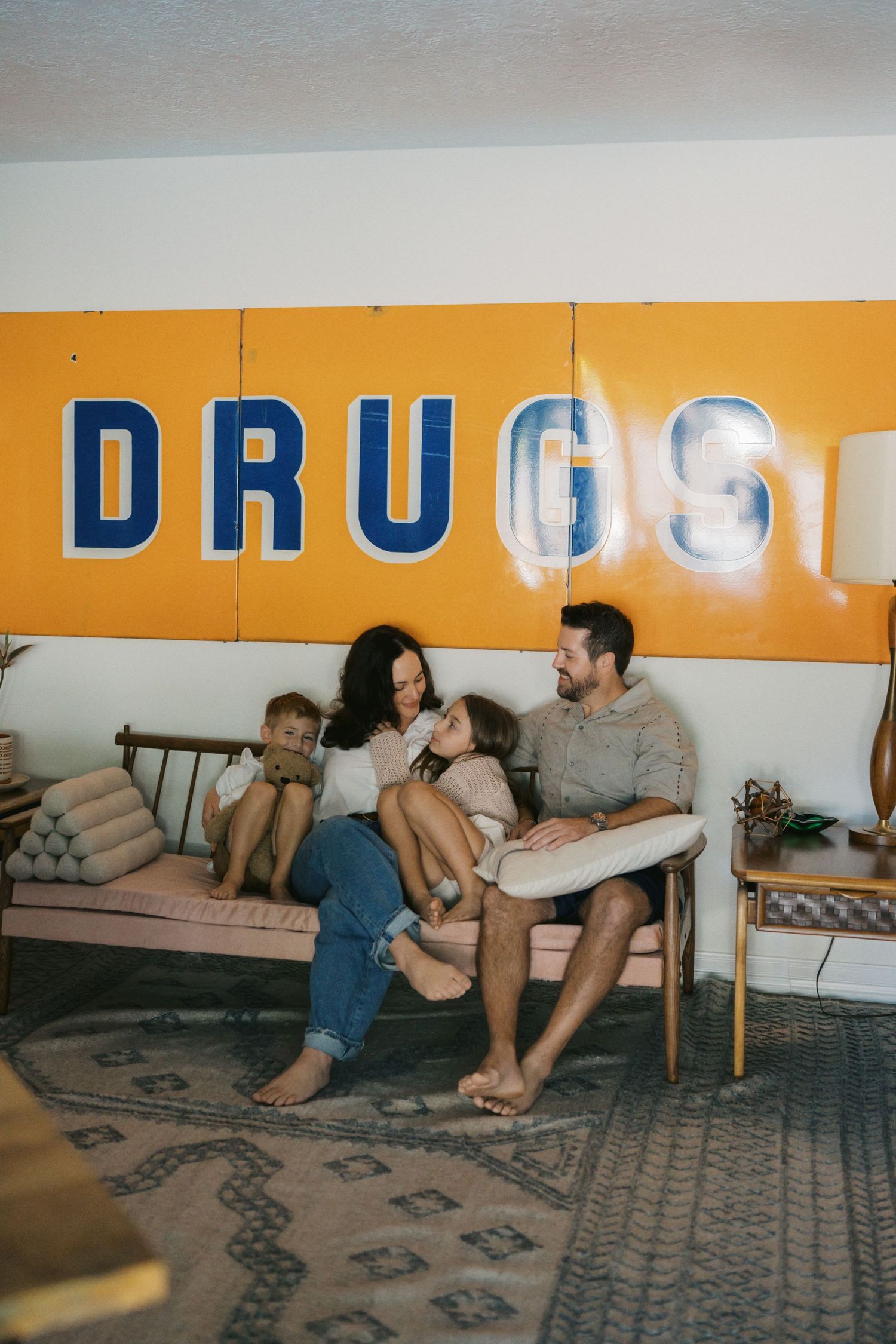 Family of four sitting on a vintage sofa beneath a retro yellow DRUGS sign in a mid-century modern living room.