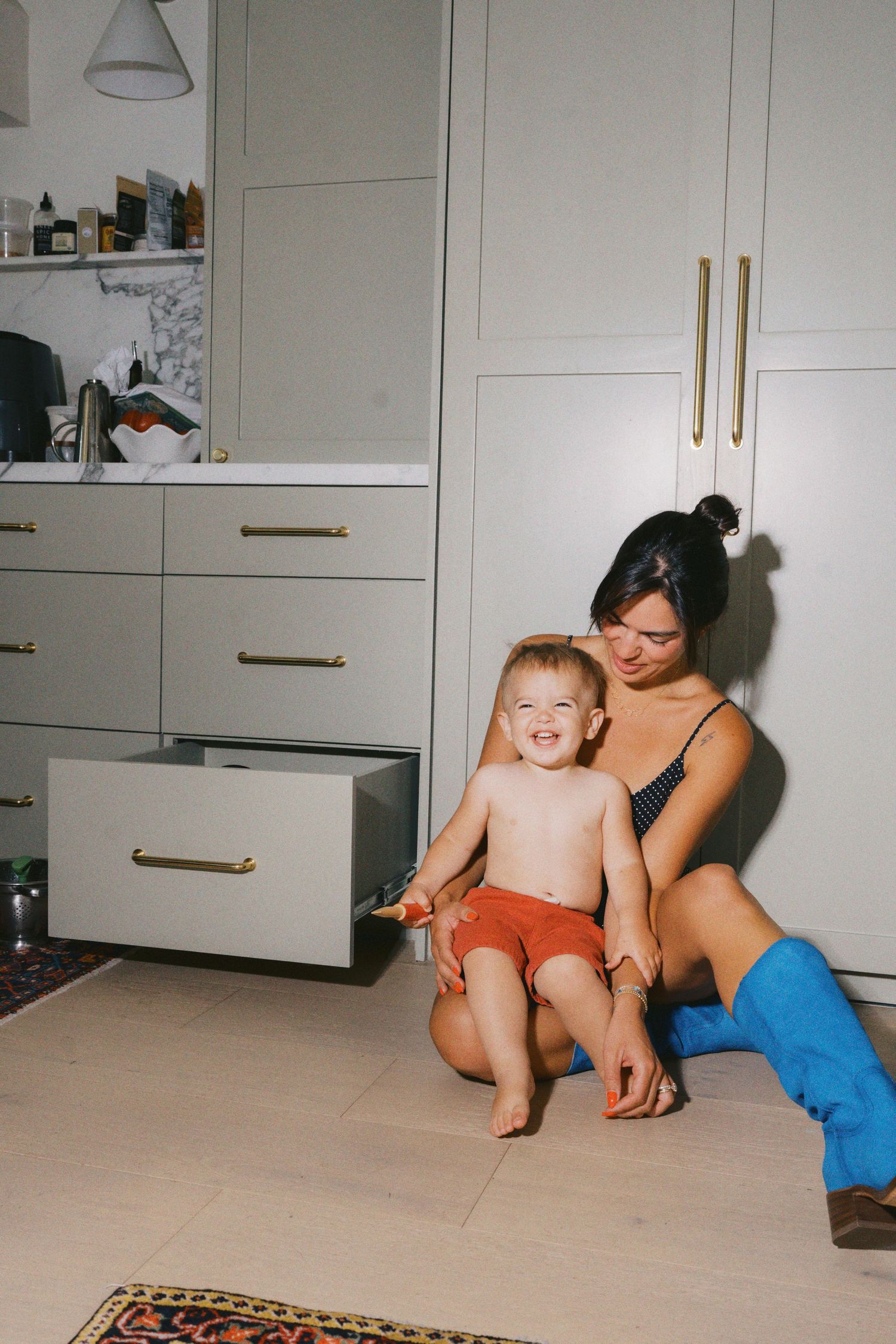A woman sits on the floor with a smiling toddler in red shorts near gray cabinetry in a modern home.