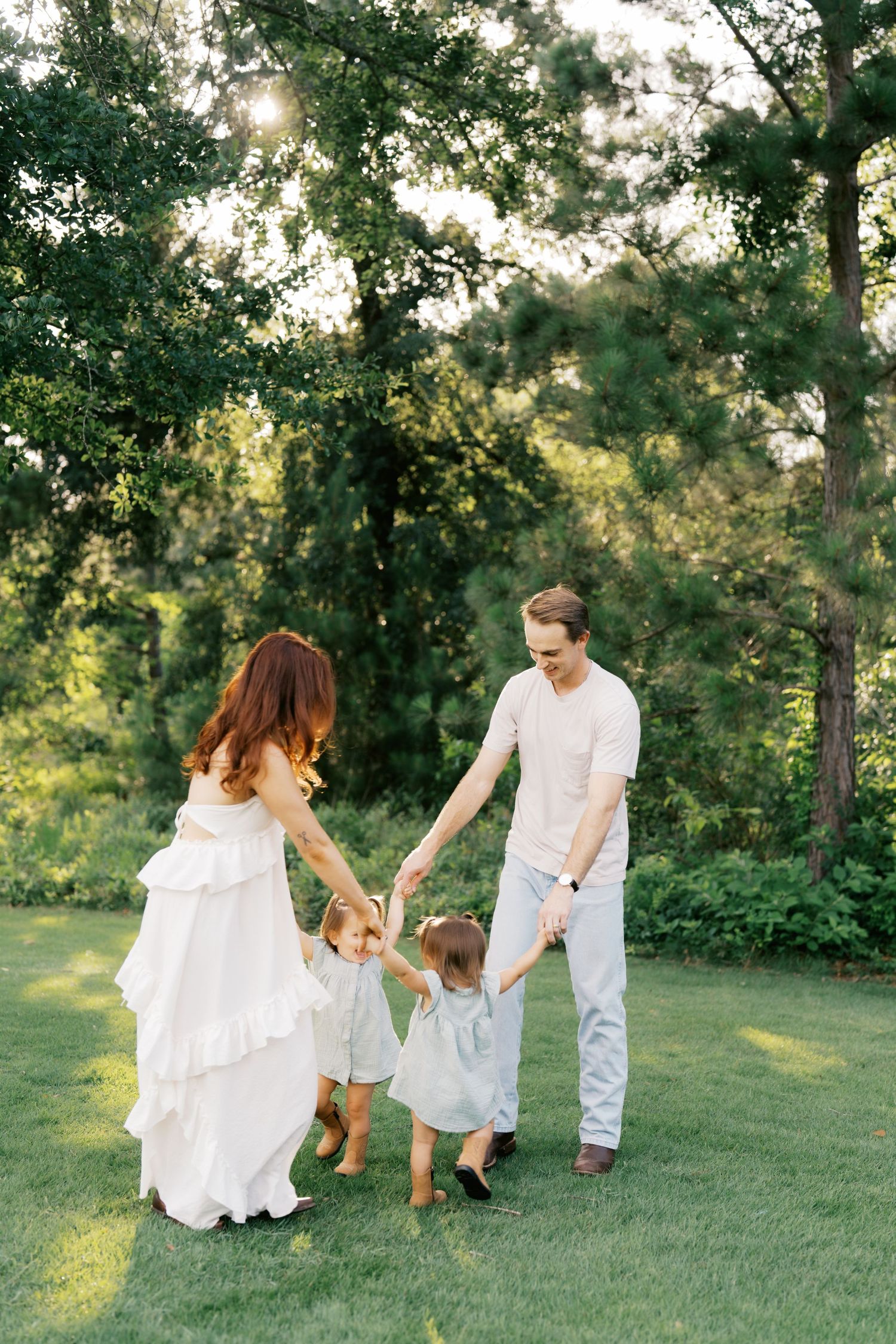 Family of four holding hands and playing in a sunlit green park, mother in white dress, father in white shirt.
