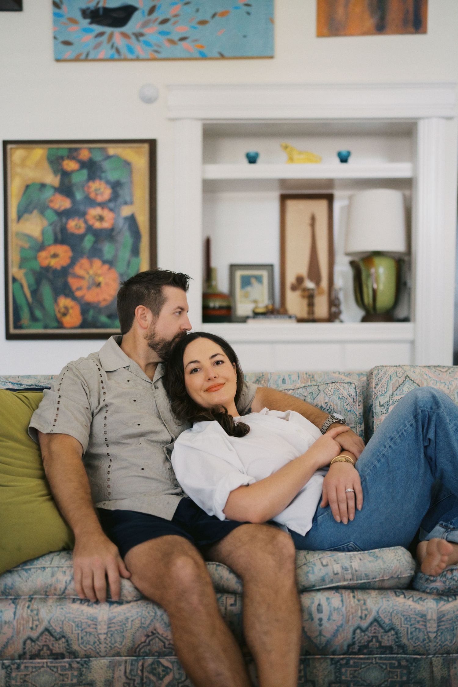 A couple relaxes on a patterned sofa surrounded by colorful artwork in a cozy, eclectic living room.