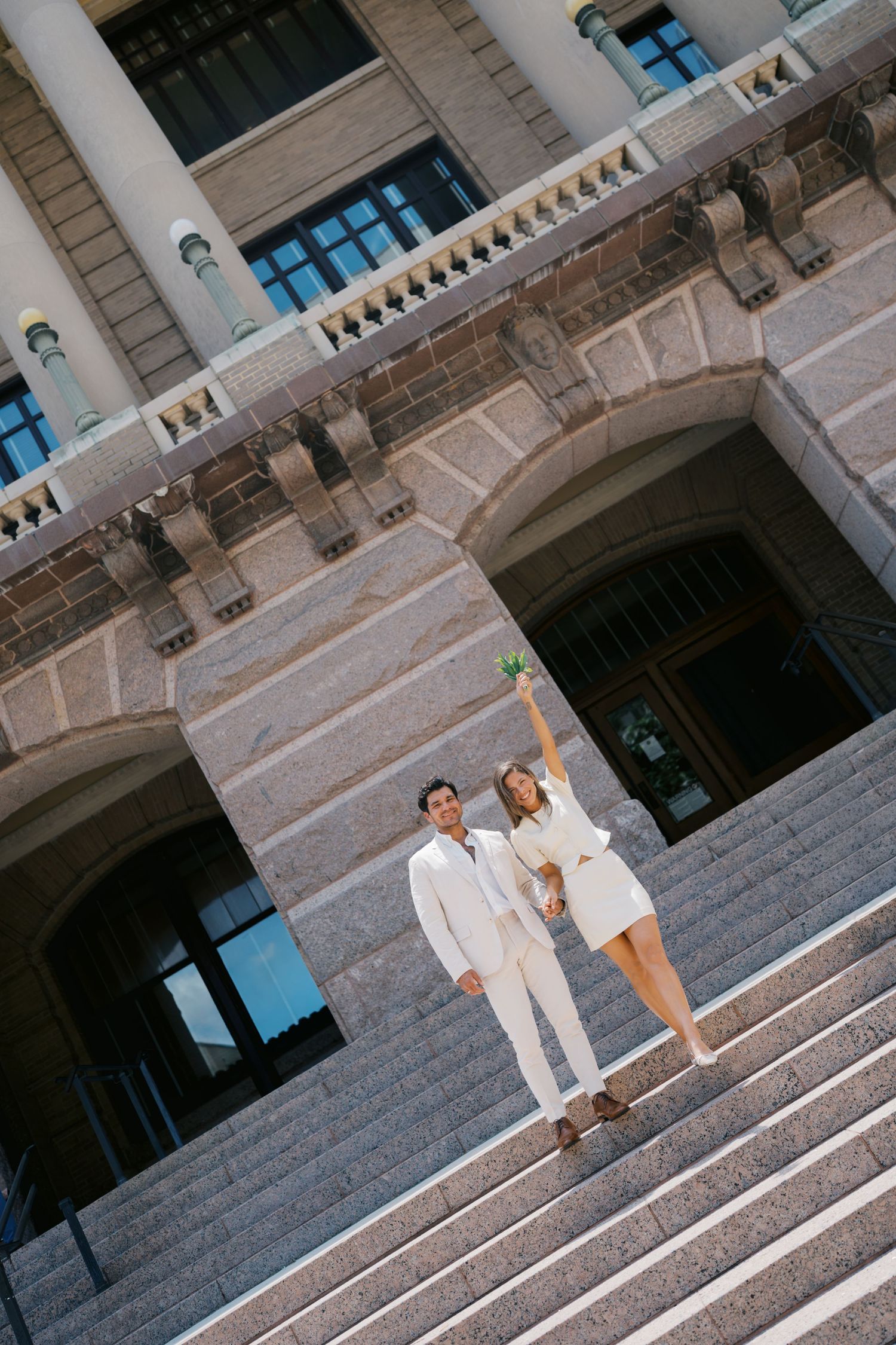 Couple in white wedding attire celebrating on grand stone steps of elegant building.