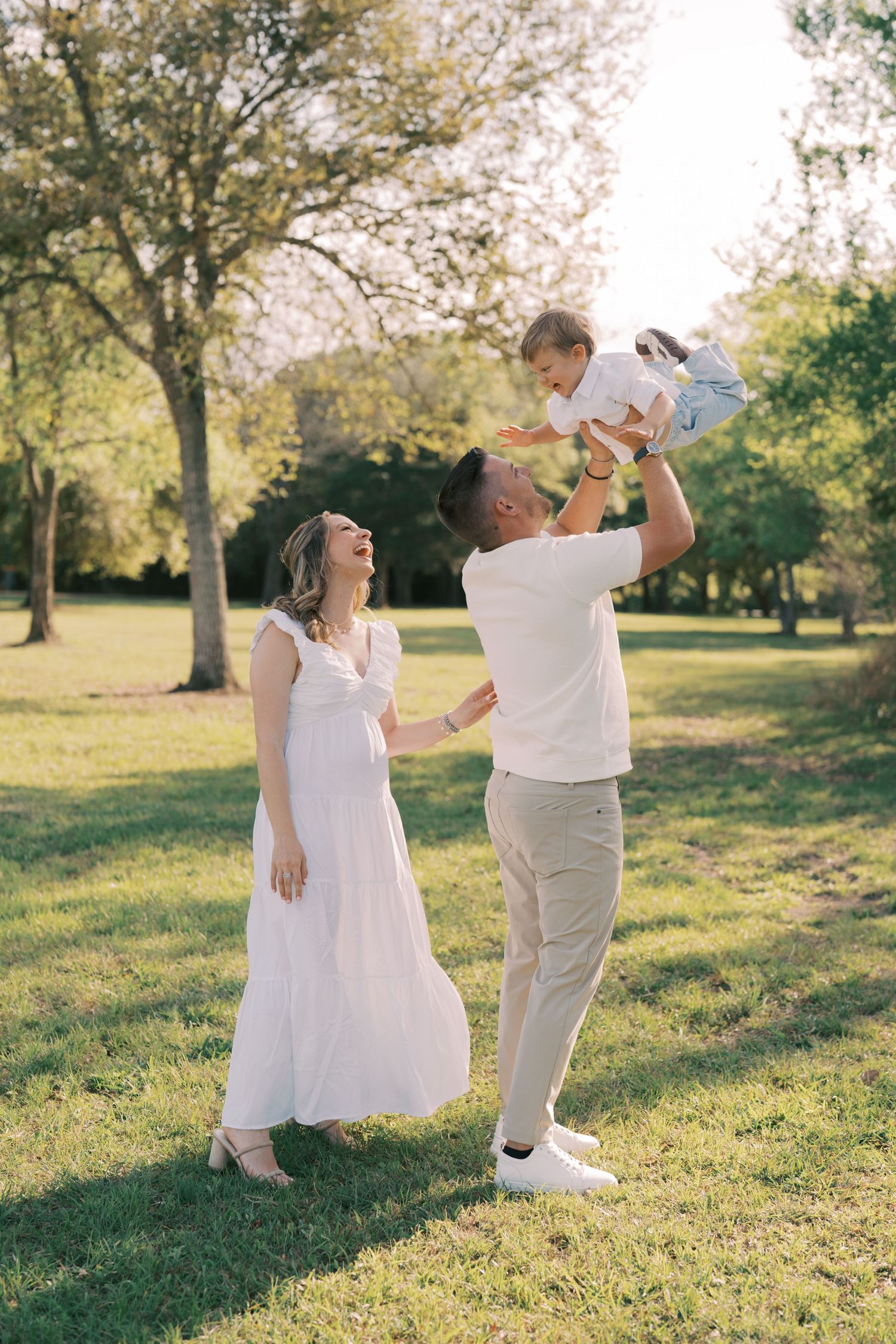 A family enjoys a tender moment together in a sunlit park with trees, wearing white and light colored clothing.