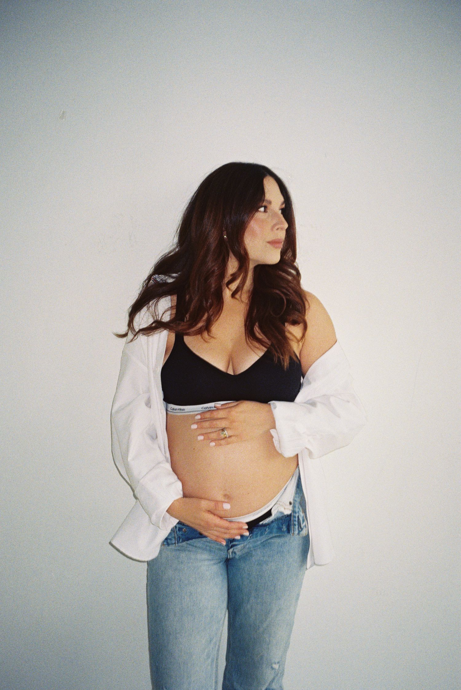 Pregnant woman in black bralette, open white shirt, and jeans cradling baby bump against white background.