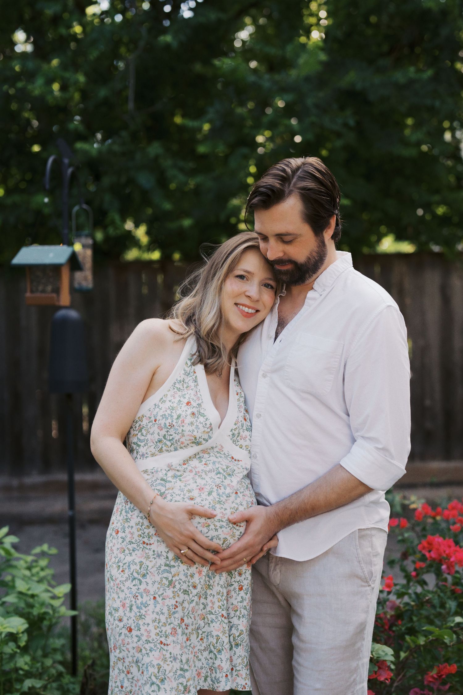 Pregnant woman in floral dress embraced by partner in backyard garden, maternity photo session outdoors.