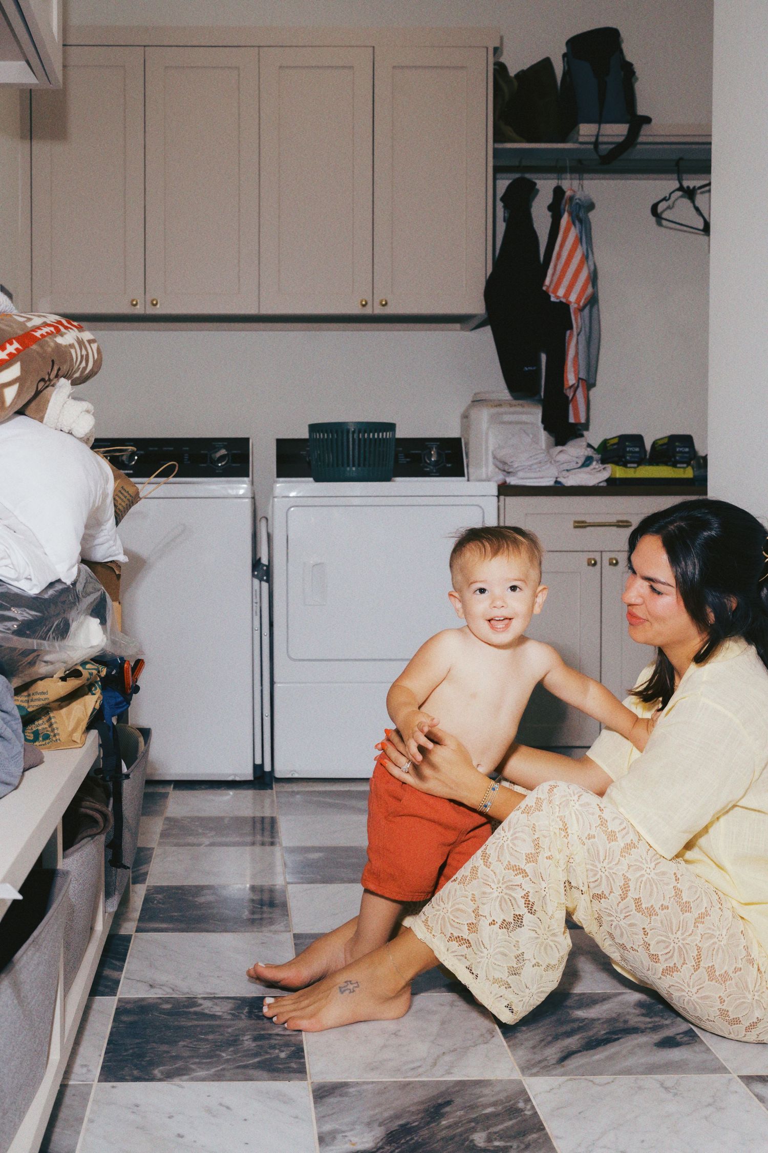 A mother sits on a laundry room floor smiling at her shirtless toddler in red shorts near a washer and dryer.