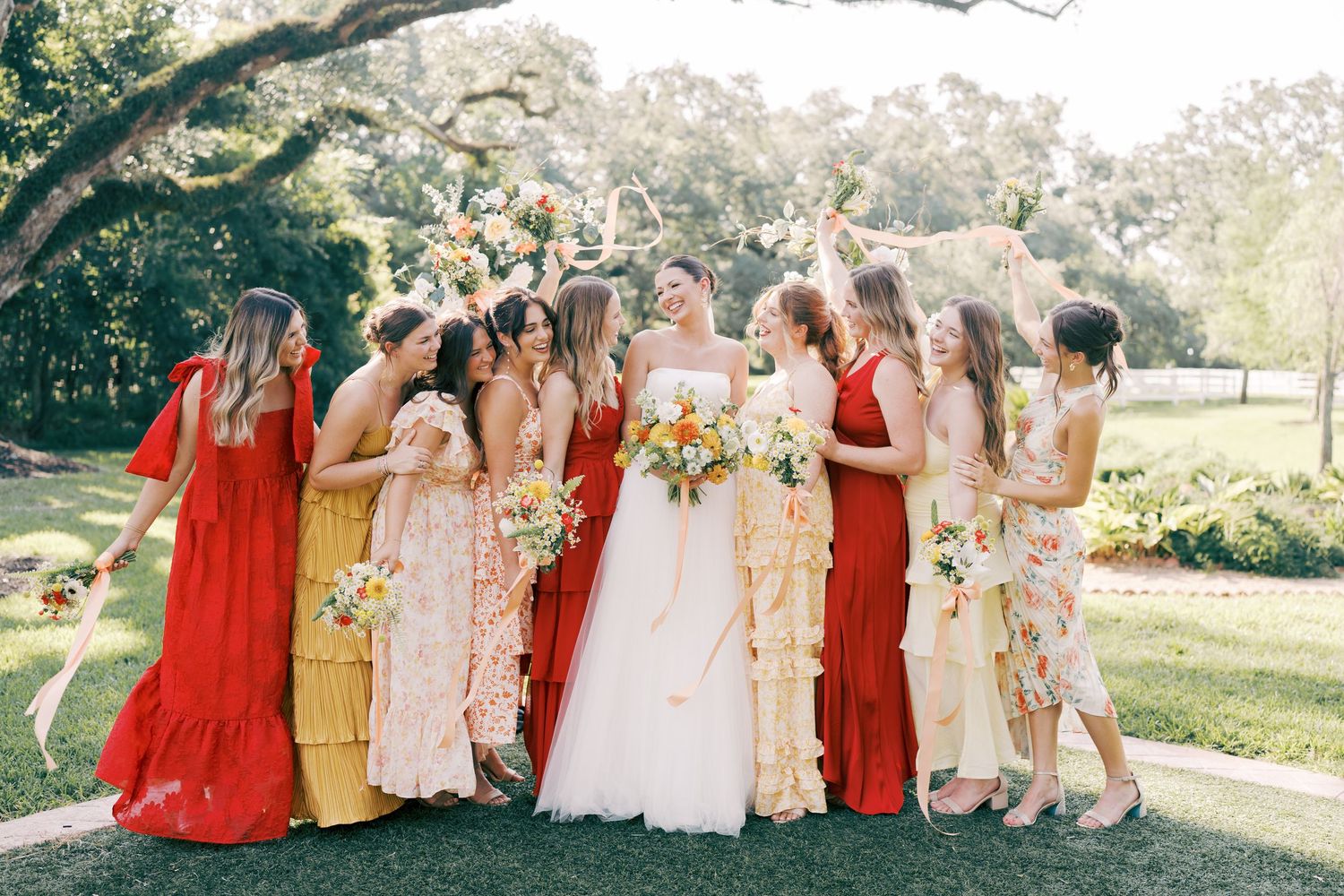A group of bridesmaids in flowing dresses of red, pink and floral patterns gather outdoors in a garden setting.